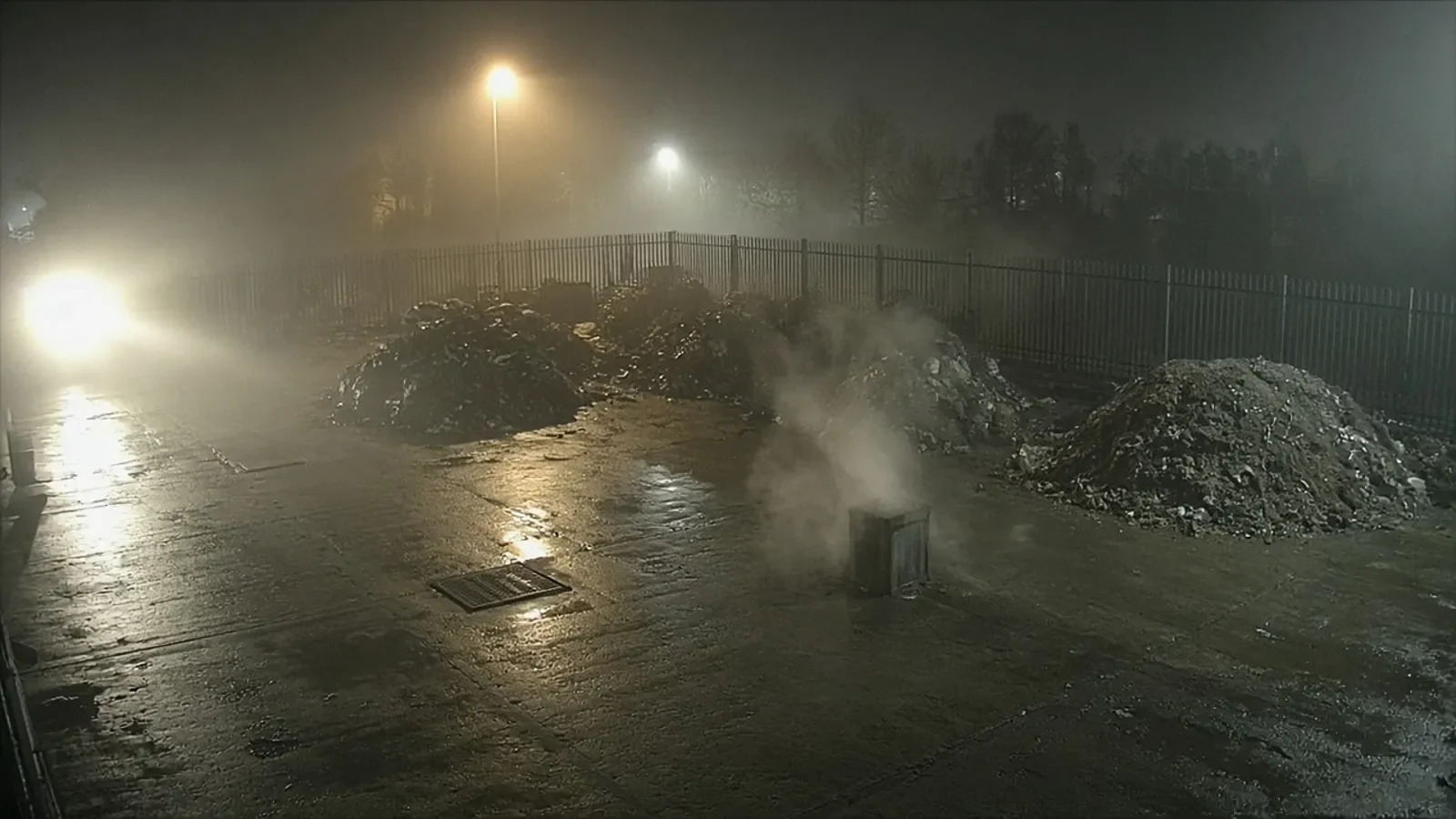 Night camera view of an empty yard in fog, with steam rising from a vent between spoil heaps and car headlights glaring through the mist
