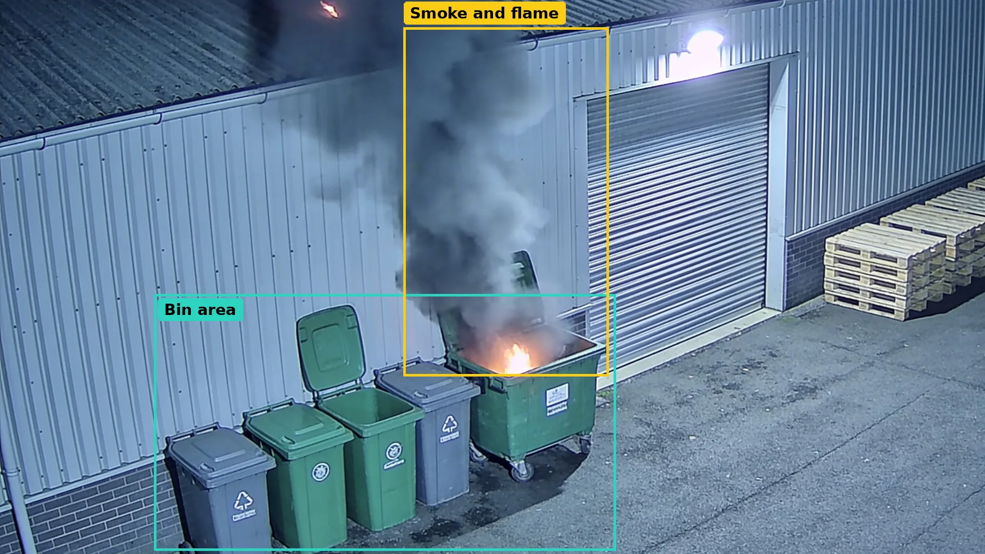 Night CCTV of a rear yard: wheelie bins against the wall in a teal zone, one alight with smoke boxed in amber