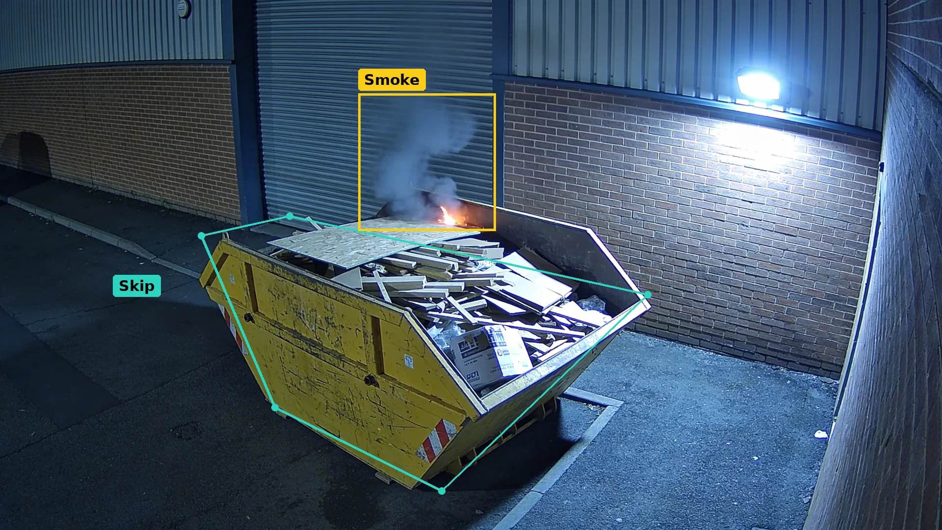 Night CCTV of a yellow skip of timber and card in a teal zone near a wall, smoke and flame boxed in amber