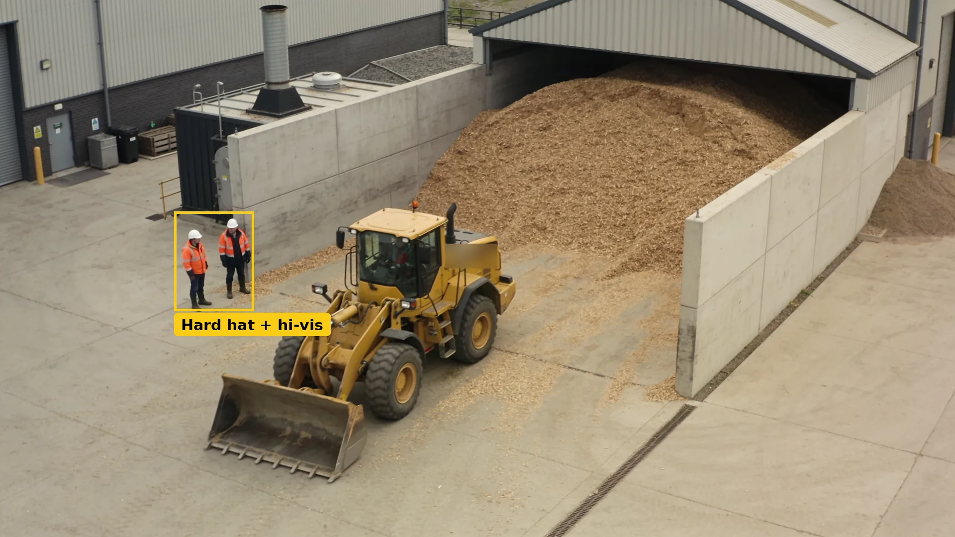 Daytime CCTV of a chip store yard: two staff in PPE boxed in teal beside a parked loading shovel