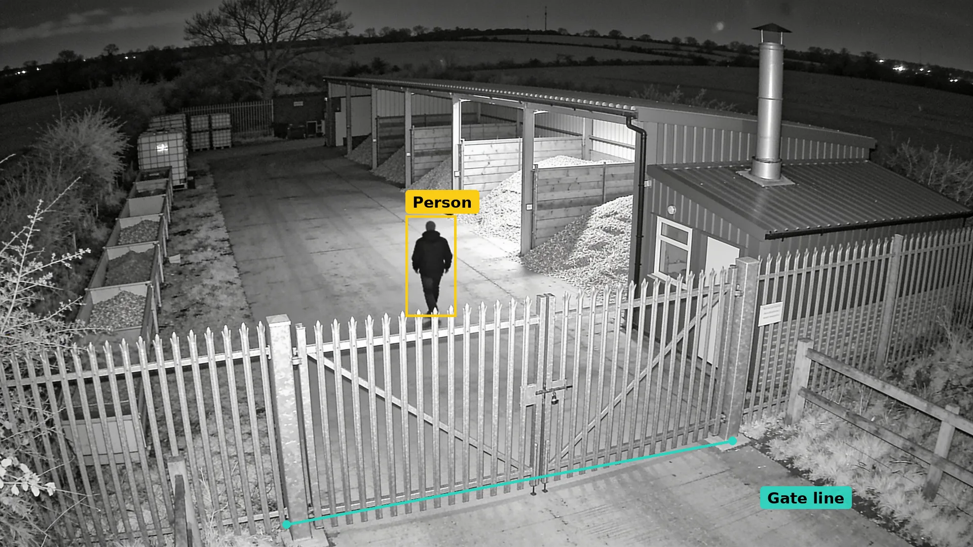 Infrared CCTV of a fuel yard at night: a teal line along the palisade gate and a person in the yard boxed in amber