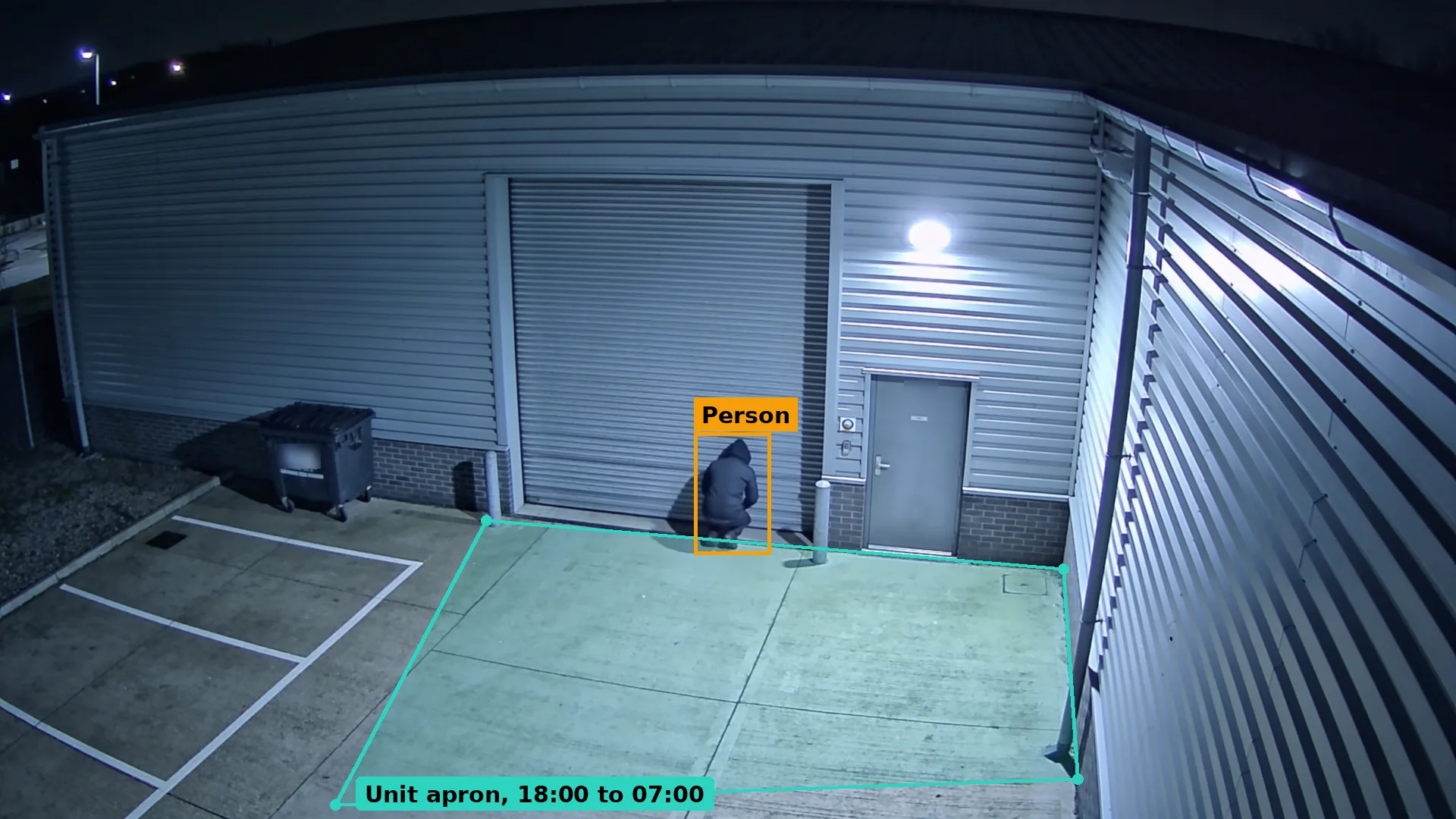 Night CCTV of a hooded person crouching at the base of a roller shutter on an industrial unit, boxed in amber