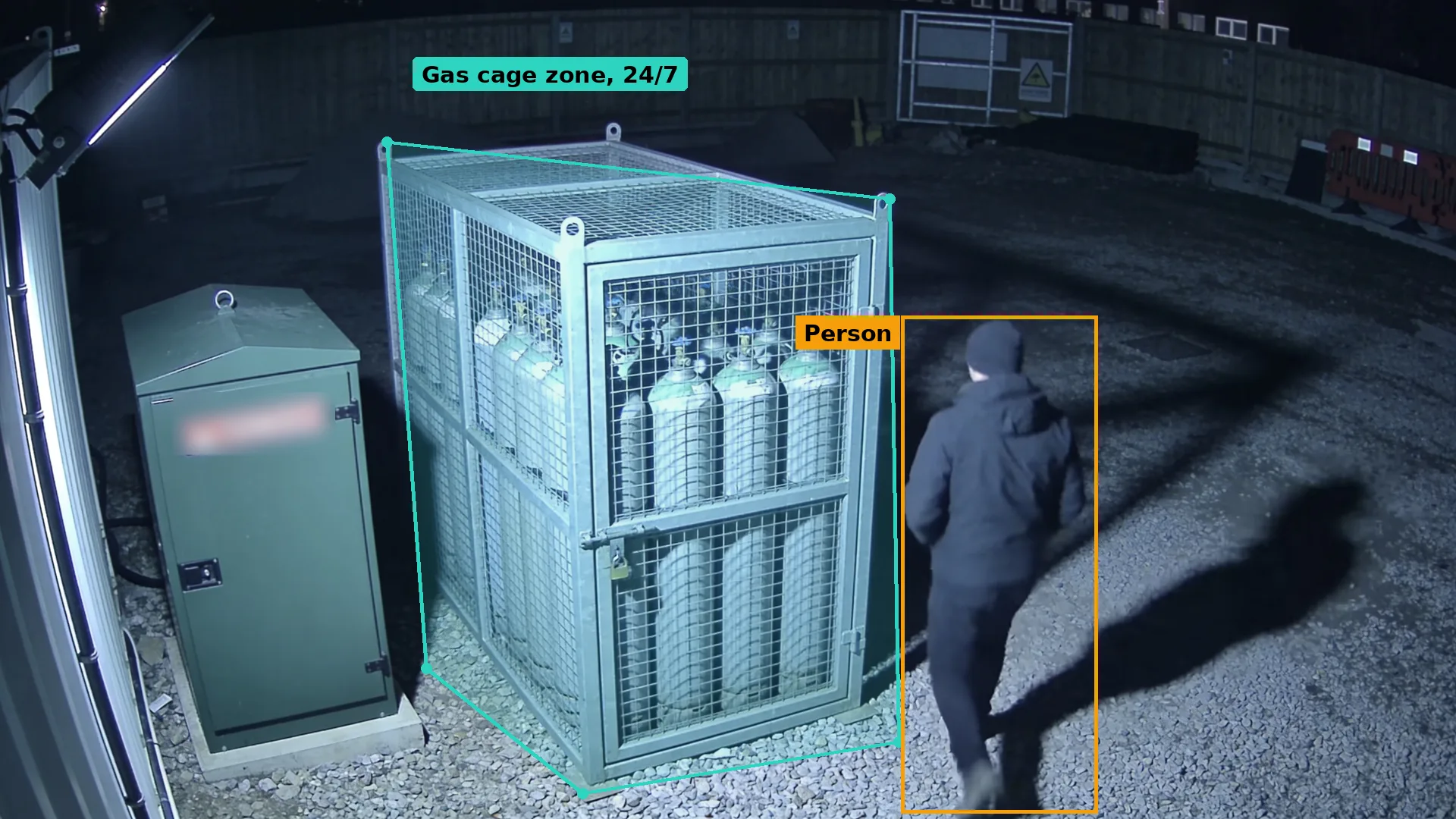 Night CCTV of a locked gas cylinder cage beside a flammables cabinet, a person approaching boxed in amber