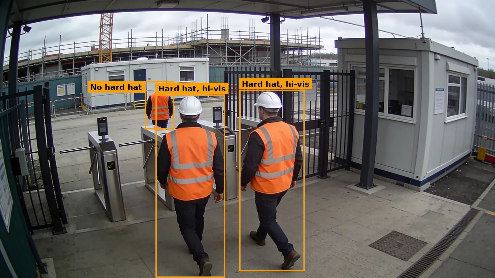 Daytime CCTV of a site entrance, two workers in hi-vis vests and white helmets, a third with no helmet, all boxed in amber