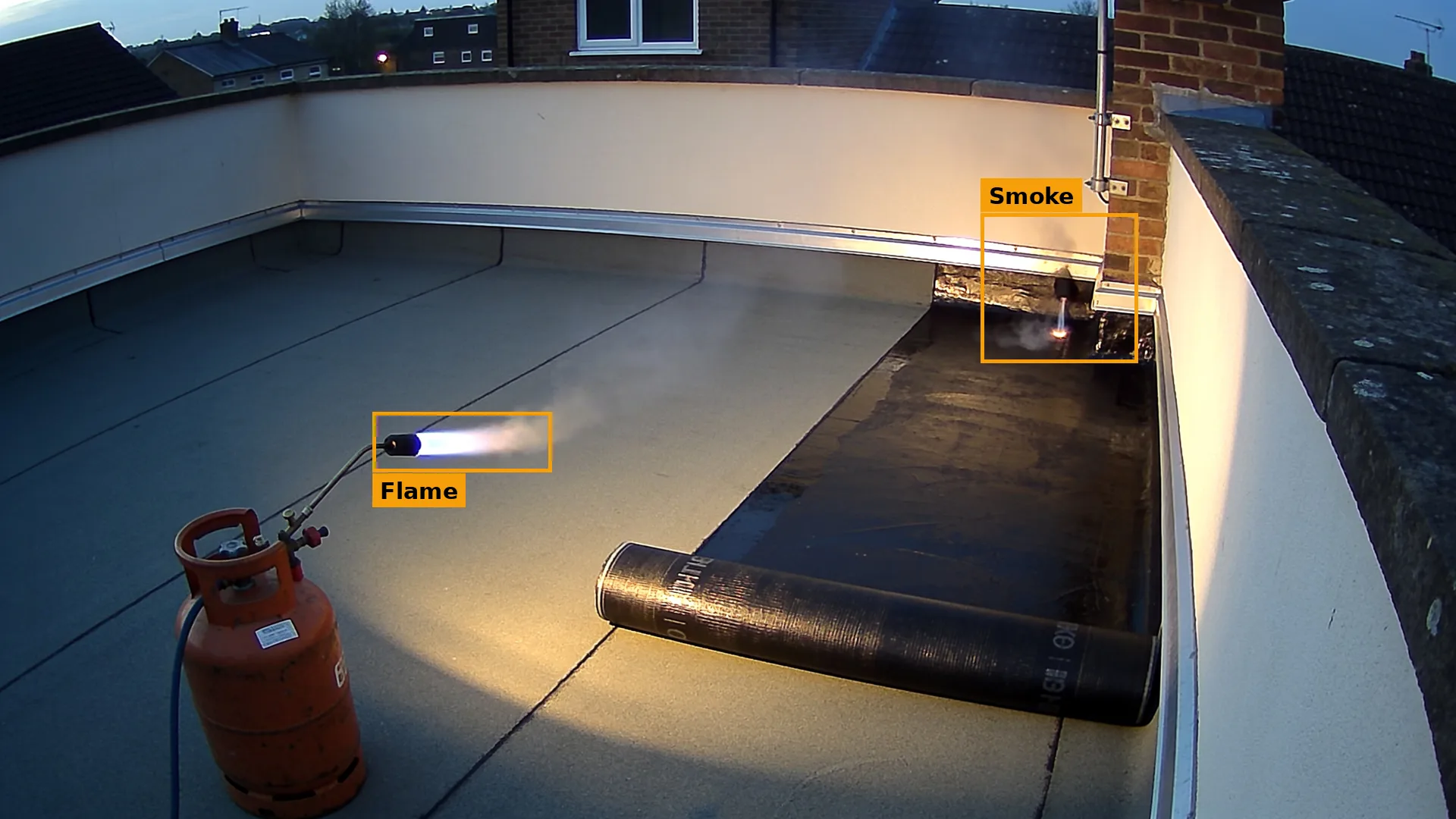Dusk CCTV of a flat roof, a lit roofing torch beside a membrane roll and smoke at the corner, both boxed in amber