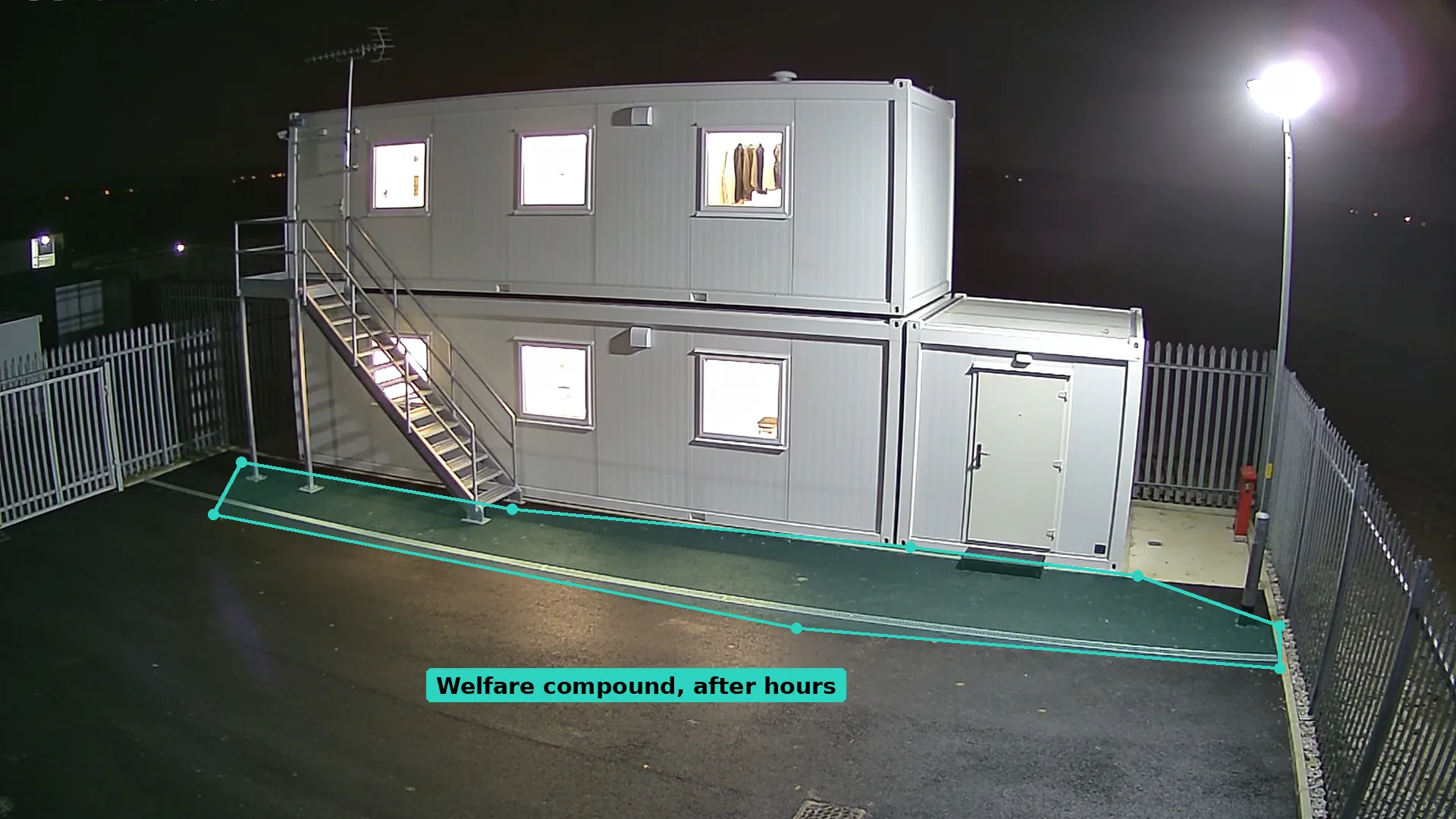 Night CCTV of stacked white site cabins with steps and a drying room, the ground in front outlined by a teal zone