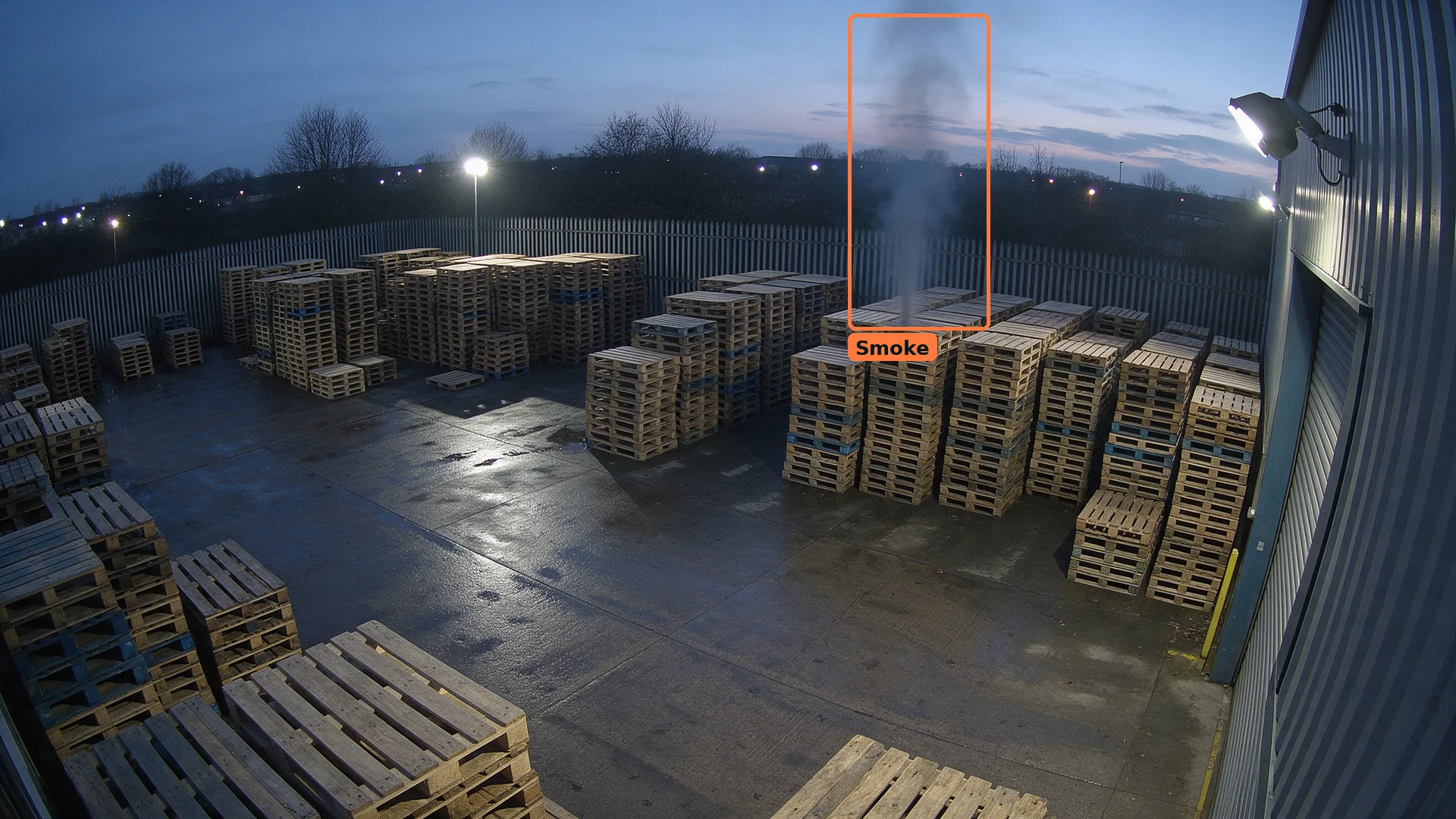 Dusk CCTV view over stacked wooden pallets in a storage yard, with a thin smoke plume boxed in orange above one stack