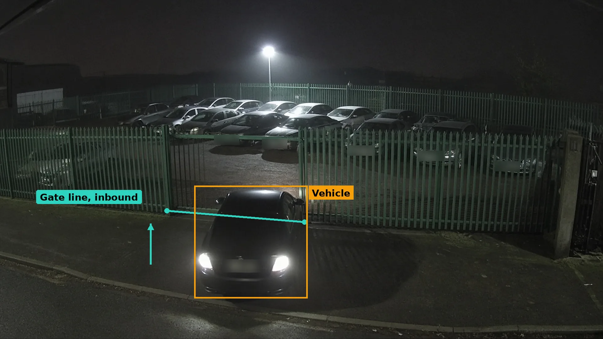 Night CCTV of a car with headlights on outside a dealership’s closed rear compound gate, a teal gate line, the car boxed