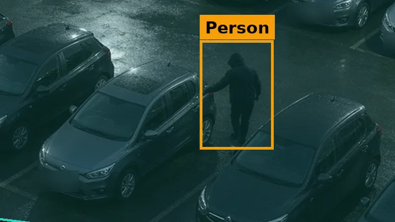 Phone alert crop: a hooded figure at a car door on a dealership forecourt at night, boxed in amber