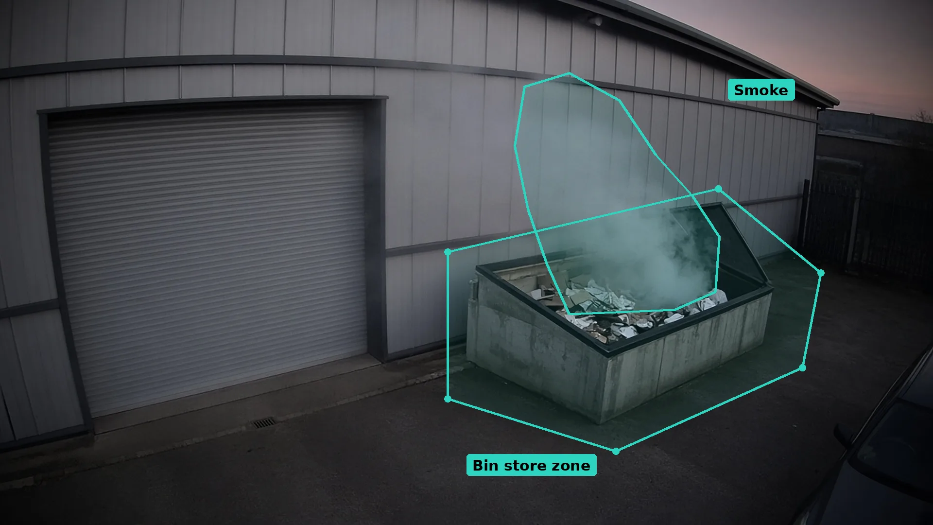 Dusk CCTV of smoke rising from an open cardboard waste bin by a dealership workshop wall, the smoke outlined in teal
