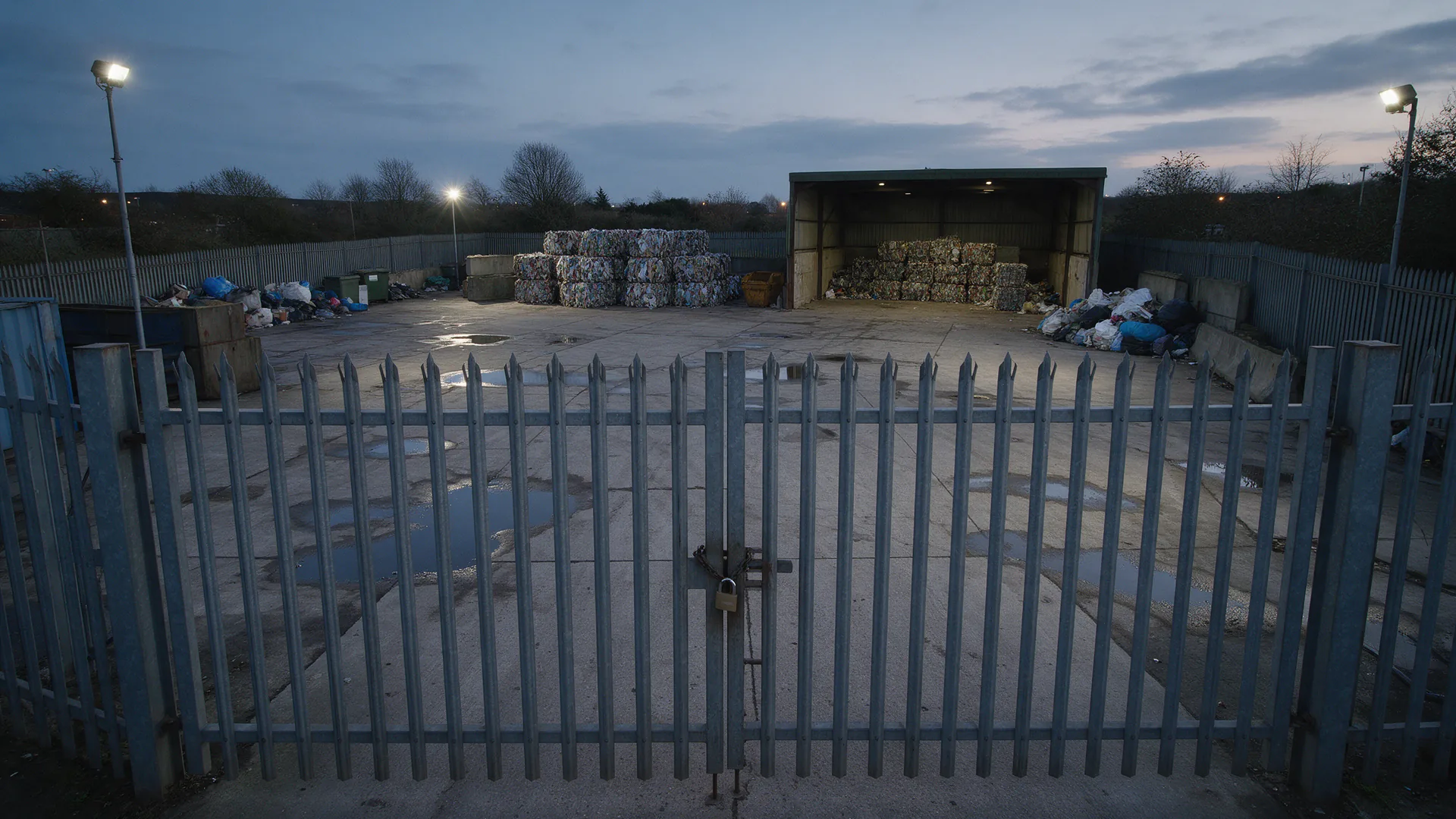 Locked palisade gate in front of an empty waste yard at dusk, with baled stock and an open storage shed under floodlights