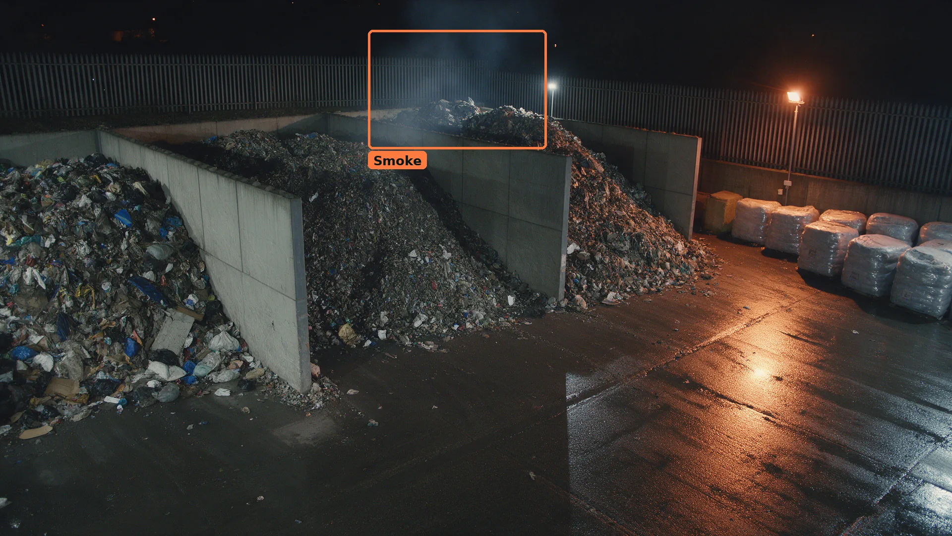 Night CCTV of three waste stockpiles in concrete bays, with smoke boxed in orange rising behind the middle pile