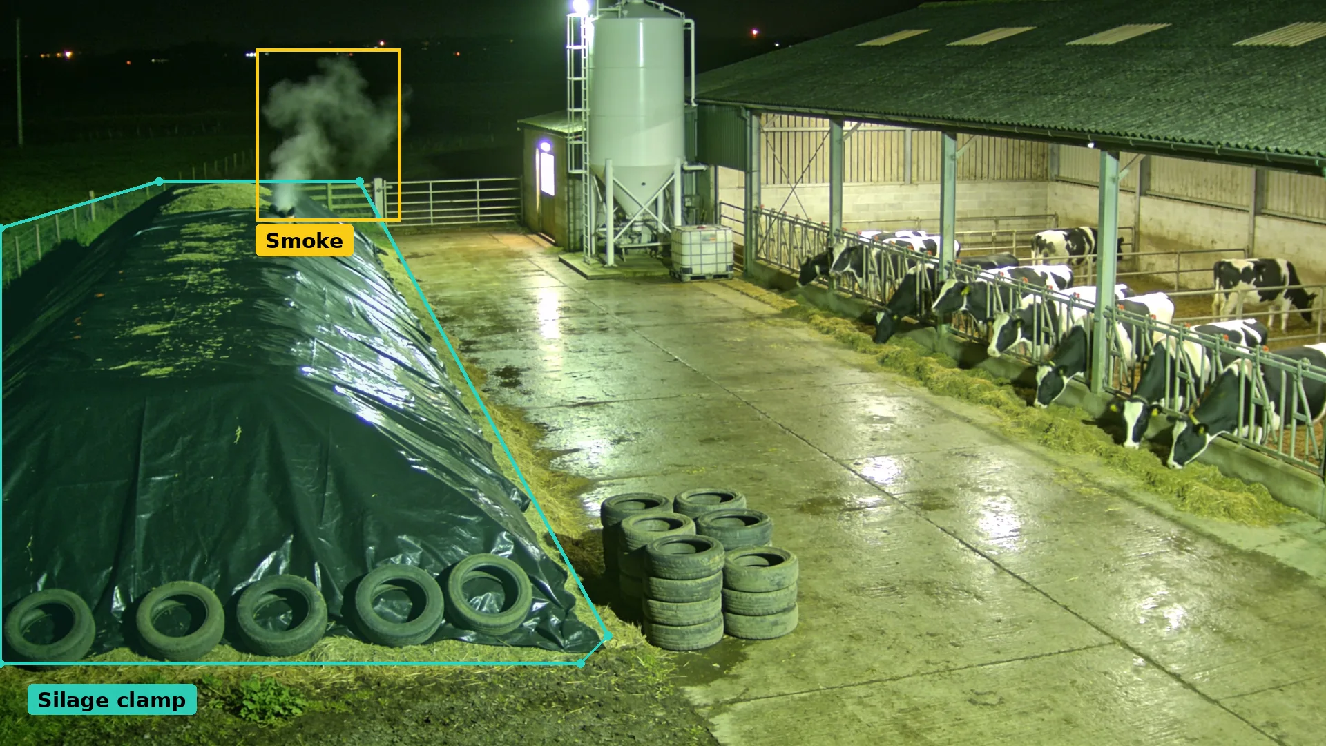 Night CCTV of a dairy yard: a sheeted silage clamp in a teal zone with smoke boxed in amber, cows feeding beyond