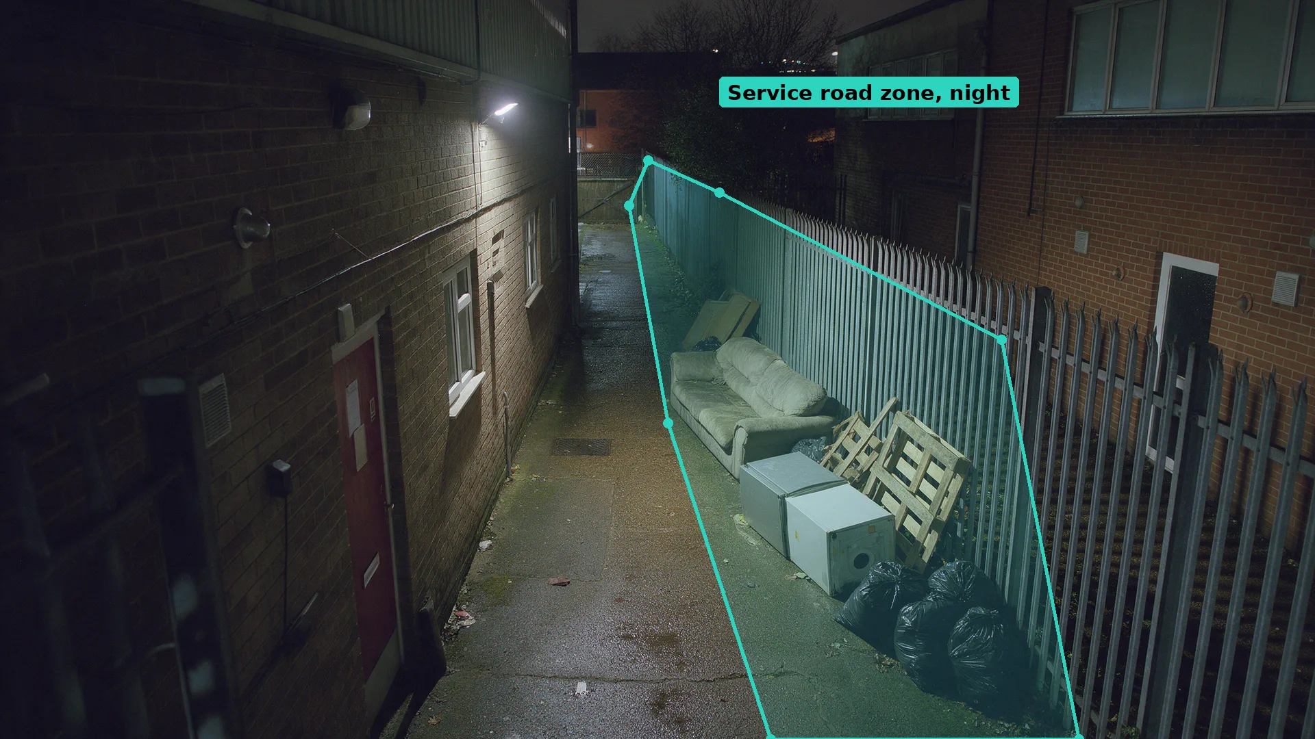 Night CCTV of a back service road with a dumped sofa, fridge and bags against the fence inside a teal zone