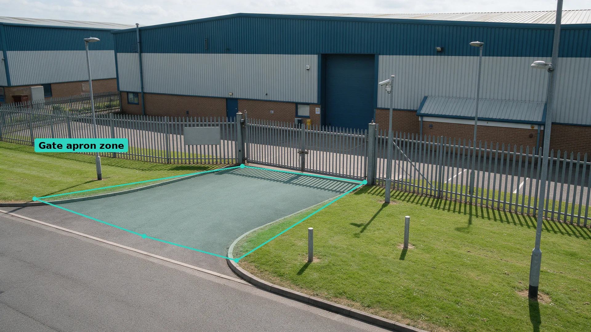Daytime CCTV of a locked palisade gate with bollards across the verge and a clean apron, the apron in a teal zone