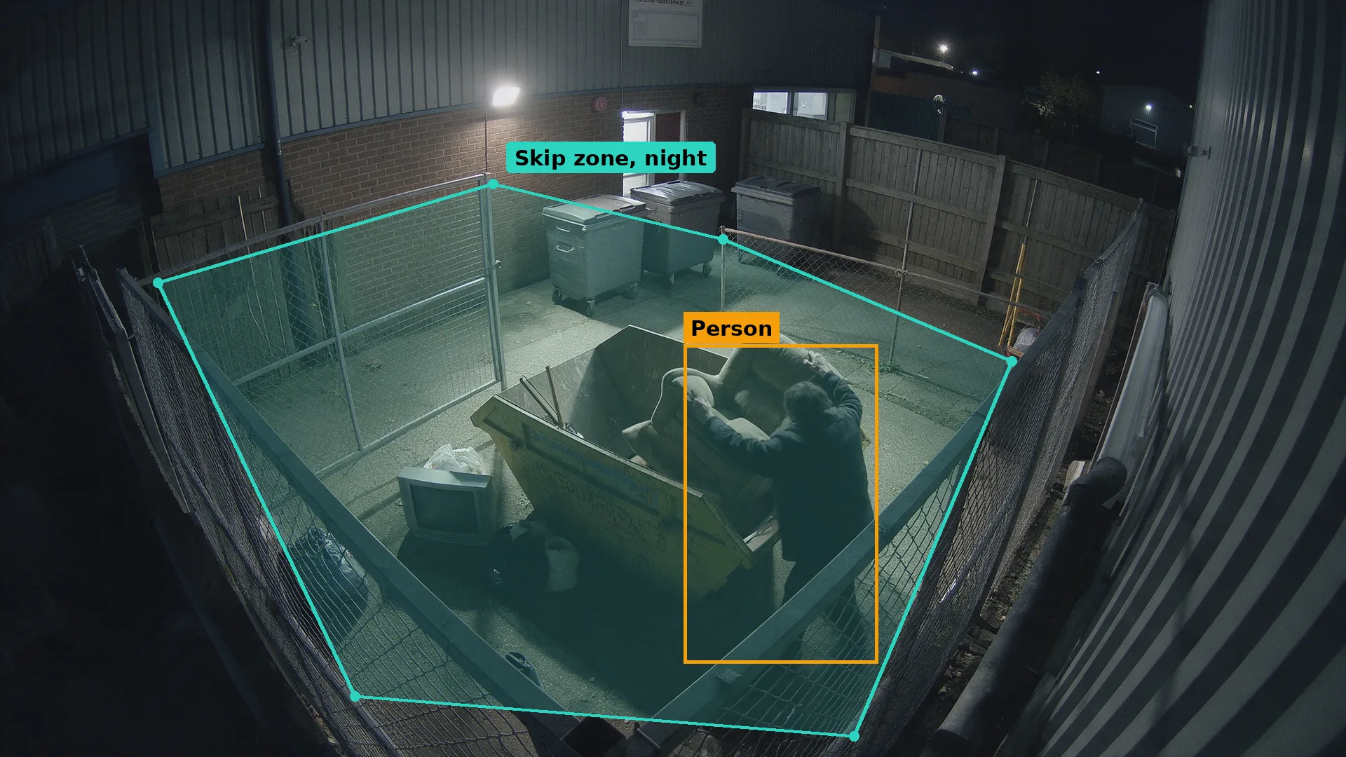 Night CCTV of a skip and wheelie bins by a wall in a teal zone, a person lifting an armchair boxed in amber