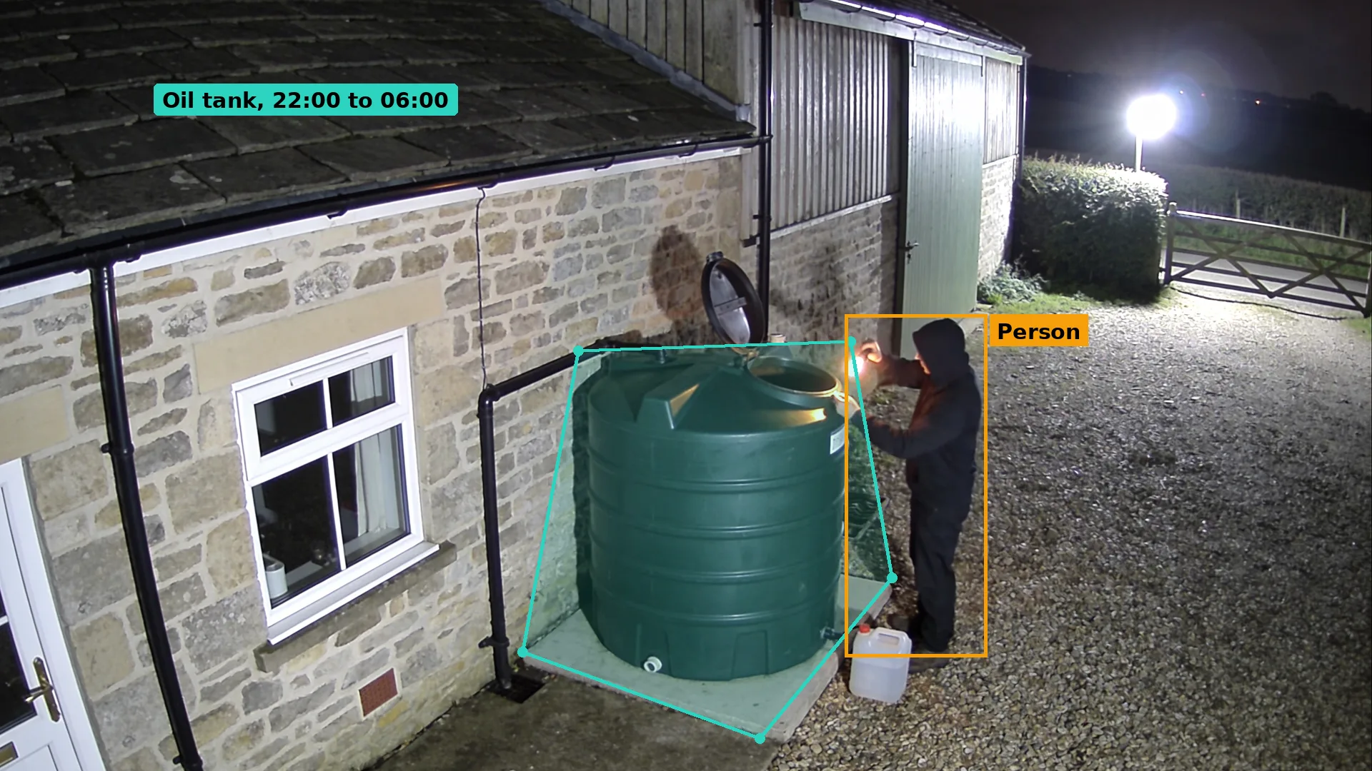 Night CCTV of a hooded person with a torch at the open lid of a green plastic oil tank beside a stone barn