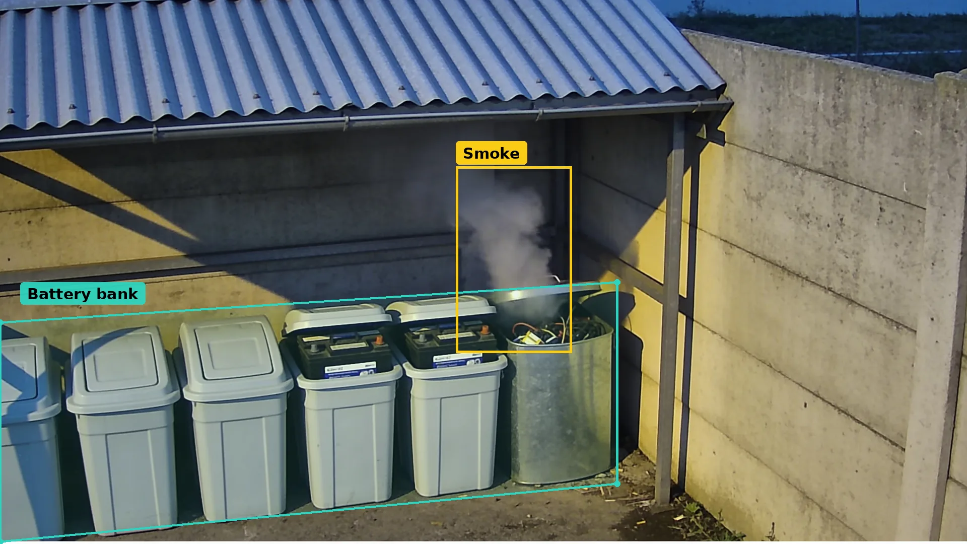 CCTV of a recycling centre battery bank at dusk: bins in a teal zone and smoke from a metal bin boxed in amber