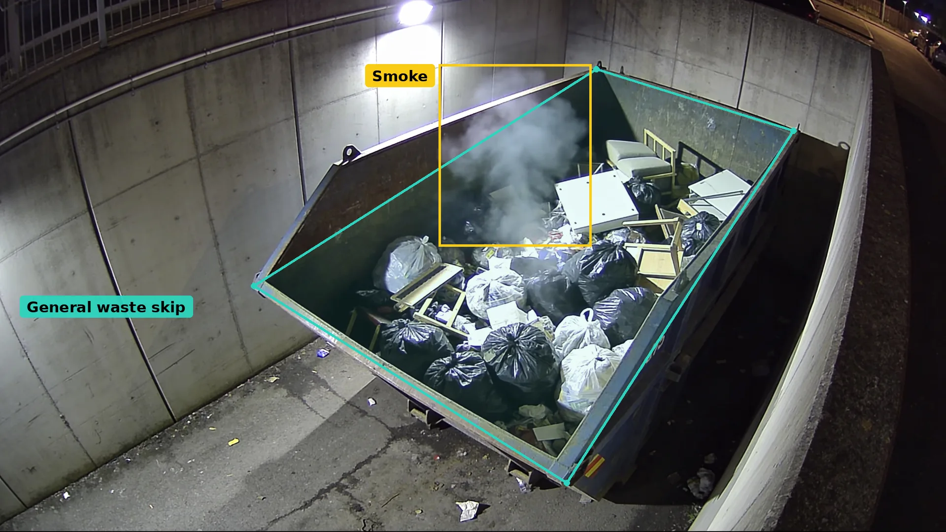 Night CCTV looking down on a general waste skip in a teal zone, smoke rising from the bags boxed in amber