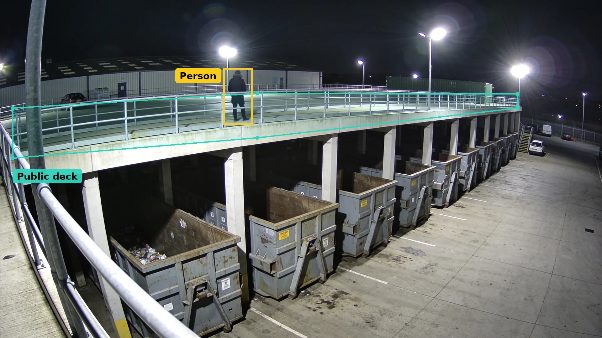 Night CCTV of a split-level recycling centre deck in a teal zone, a person on foot boxed in amber above the skips