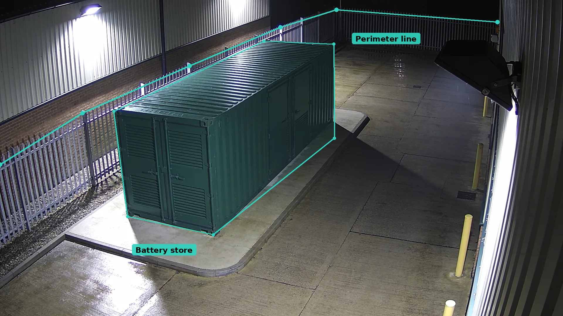 Night CCTV of a green steel container on a concrete pad in a teal zone, with a teal line along the palisade fence