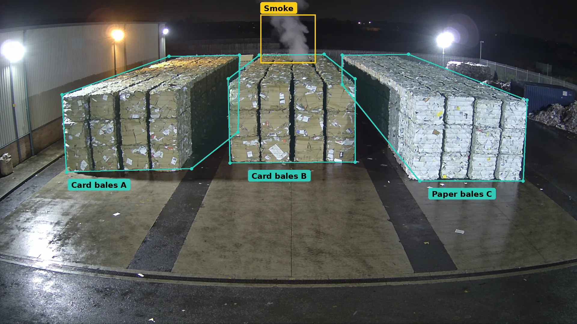 Night CCTV of three rows of paper and card bales in teal zones, with a plume boxed in amber rising from the middle row