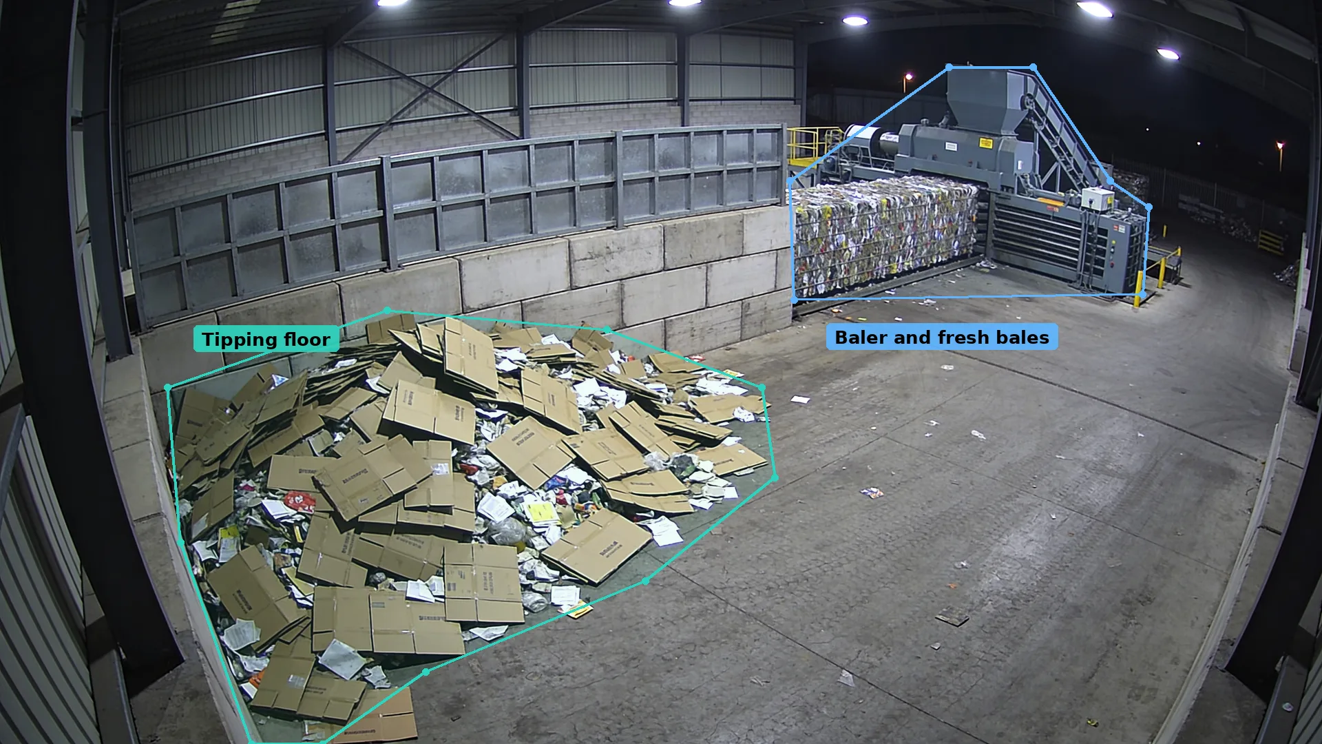 Night CCTV of loose cardboard on a shed floor in a teal zone, and a baler with fresh bales in a blue zone behind