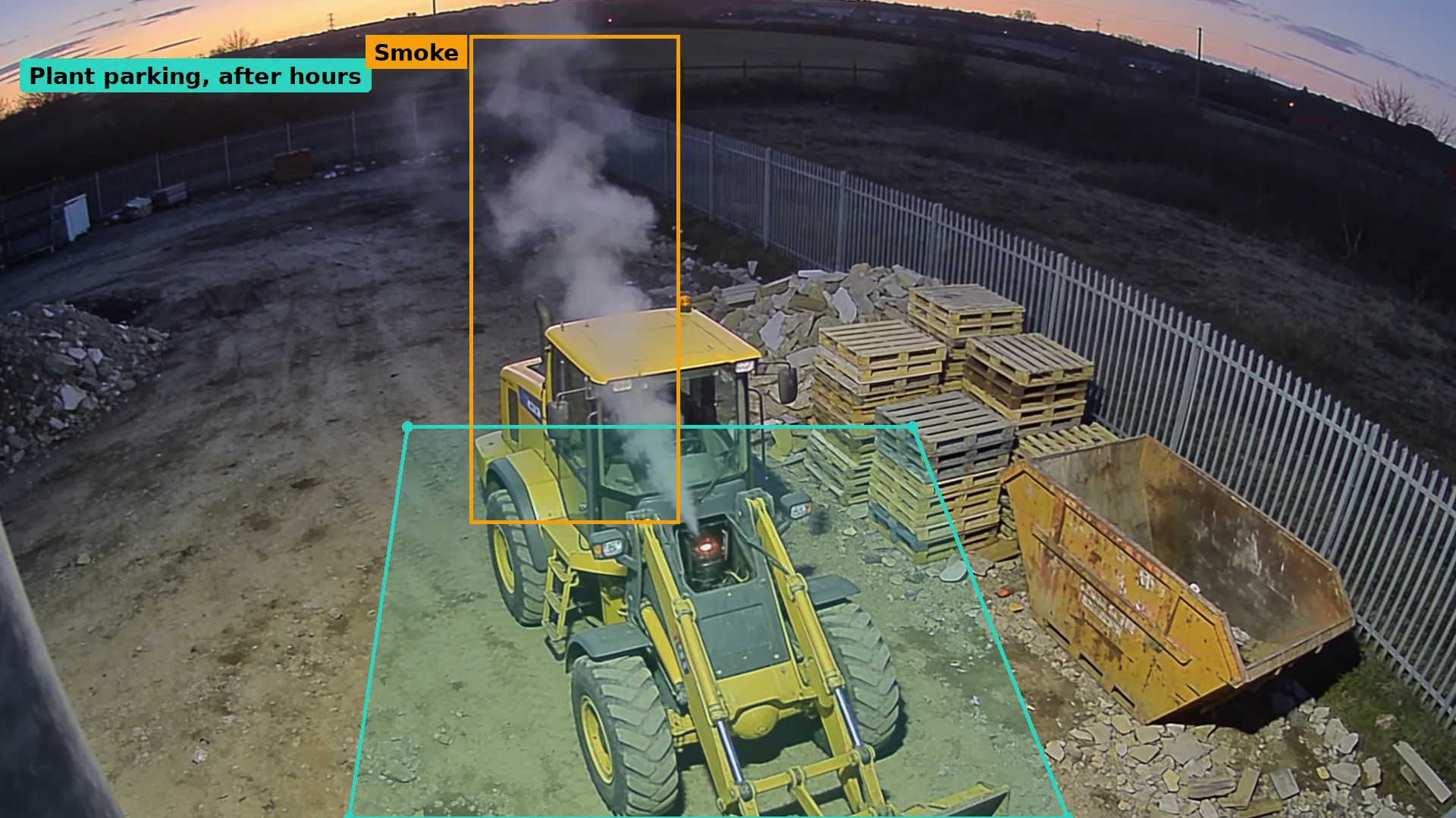 Dusk CCTV of a yellow wheel loader parked by pallets and a skip, white smoke rising from the engine bay, boxed in amber