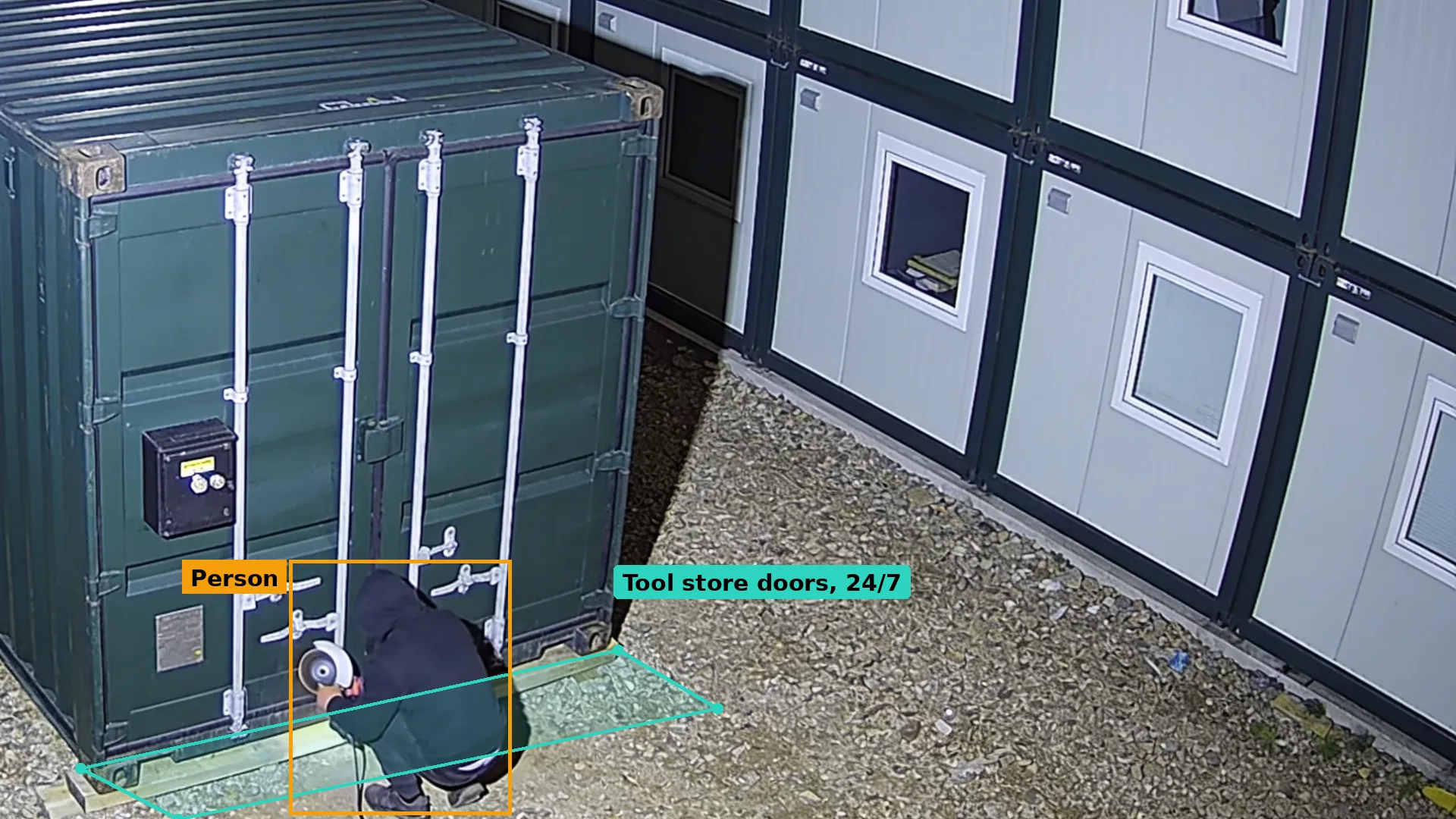 Night CCTV of a person crouching with an angle grinder at the doors of a green steel container, boxed in amber