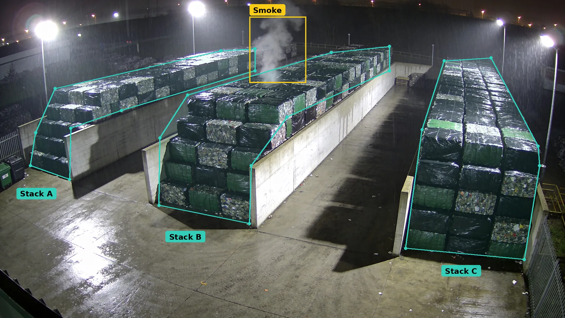 Night CCTV of a bale yard: teal zones round three stacks of wrapped fuel bales, smoke boxed in amber above the middle stack