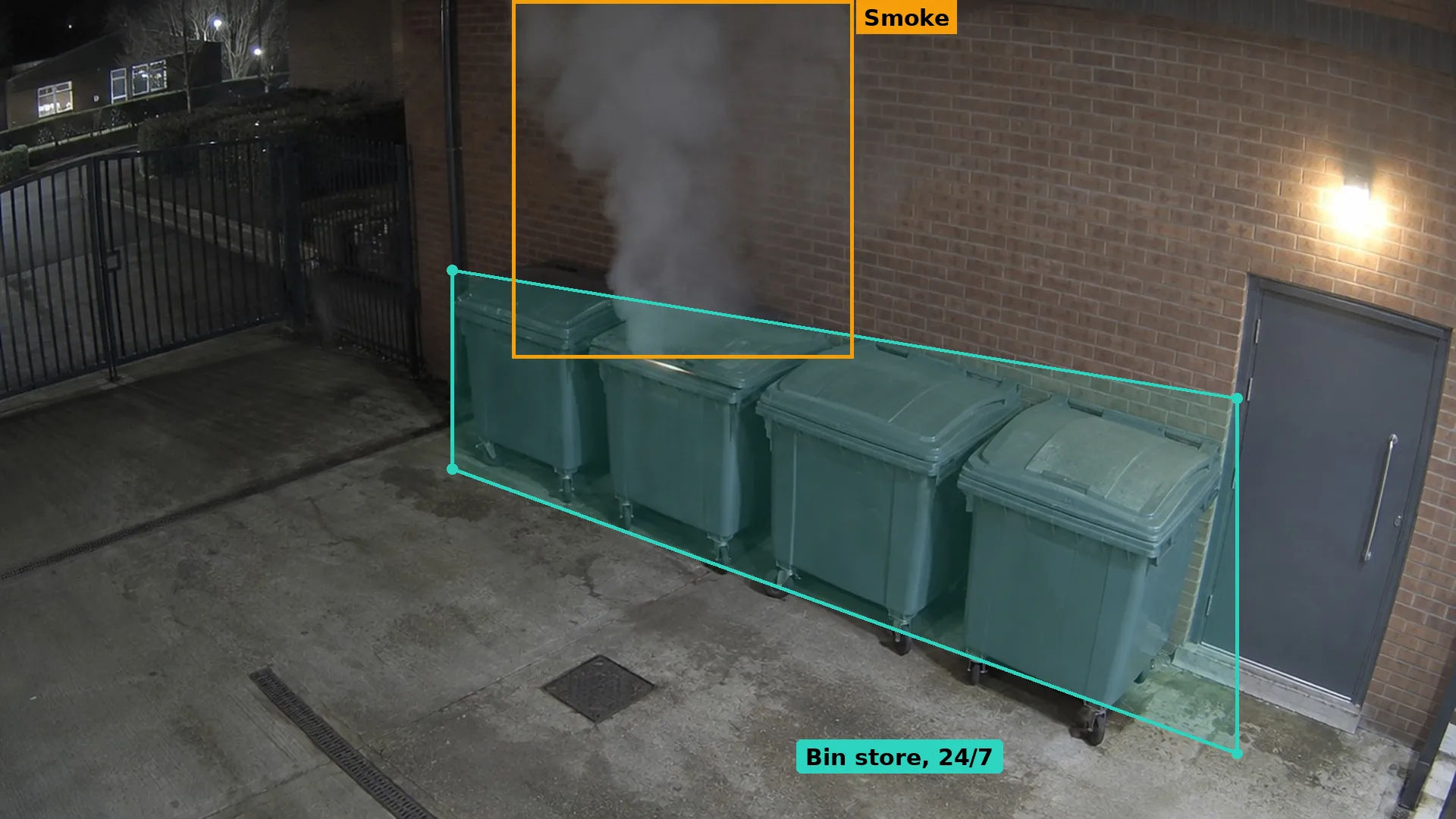 Night CCTV of smoke rising from a large wheeled bin in a school kitchen bin store against a brick wall, boxed in amber