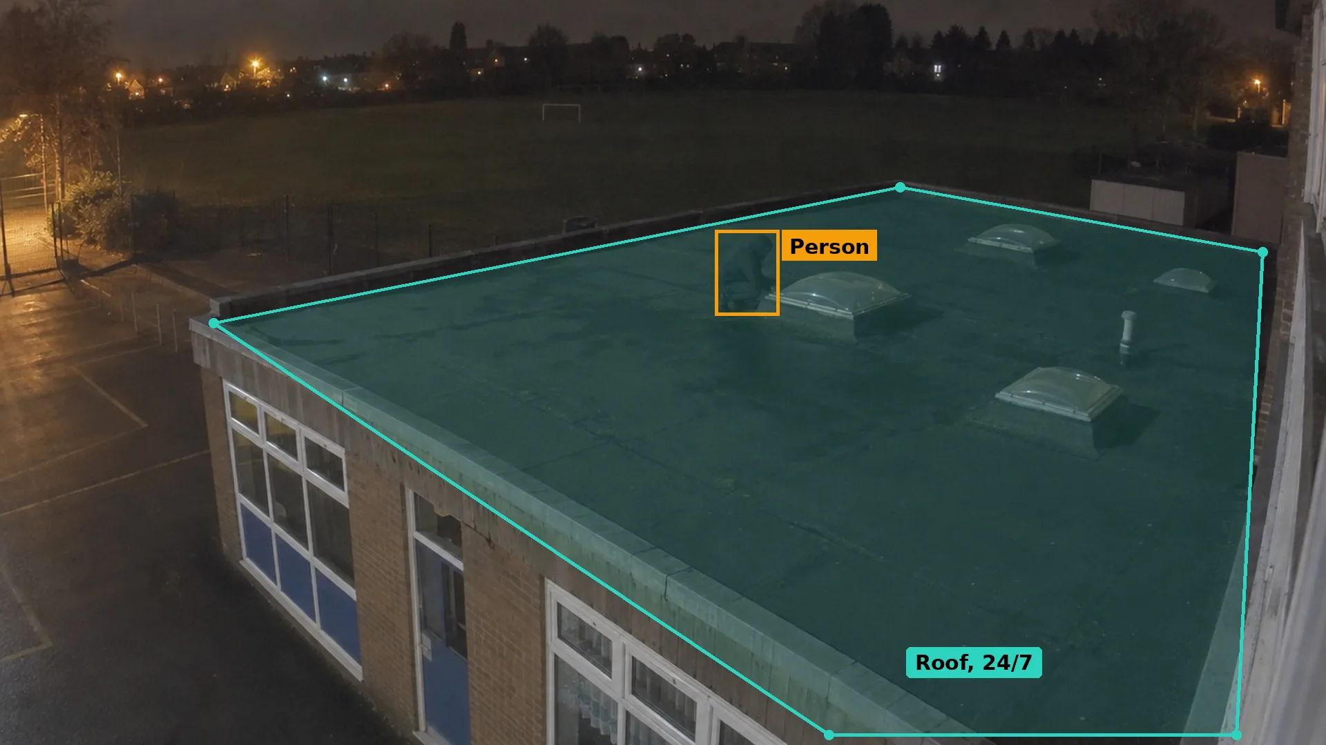 Night CCTV of a hooded person crouching beside a rooflight on the flat roof of a school classroom block, boxed in amber