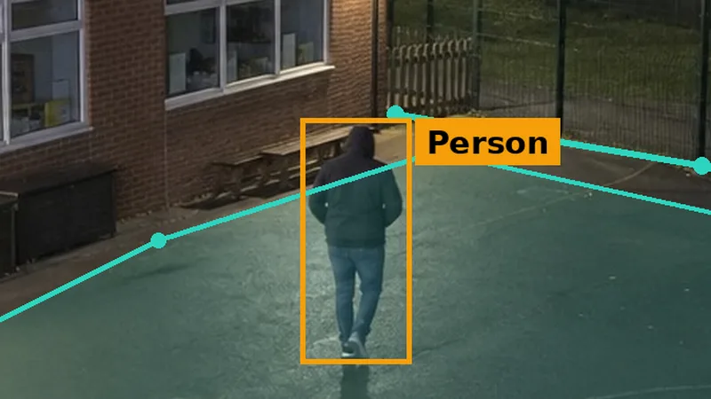 Night CCTV of a school playground: a hooded figure seen from behind, boxed inside a teal detection zone