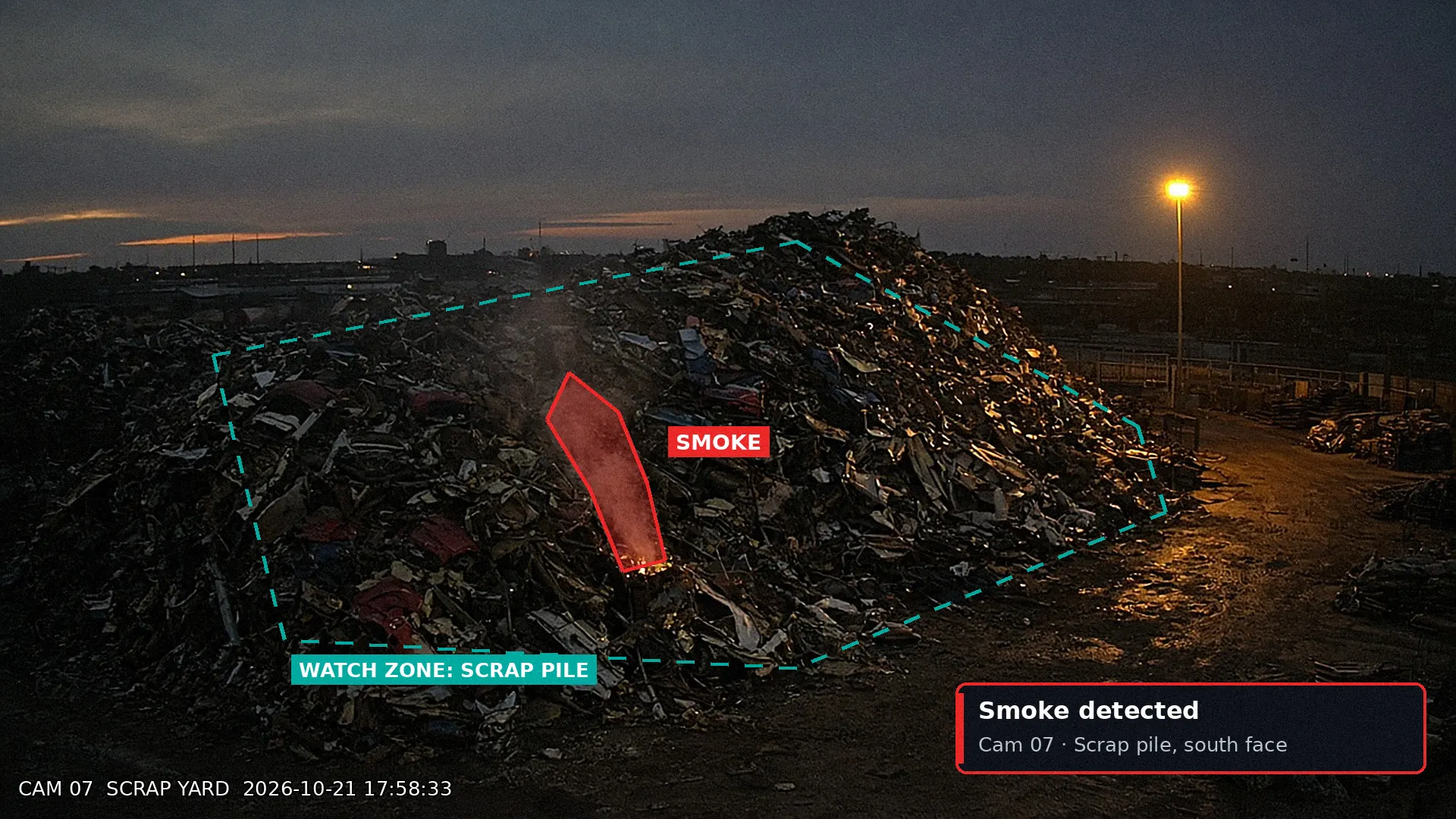 Dusk CCTV of a scrap pile with a thin smoke plume detected inside a dashed watch zone, with an alert card