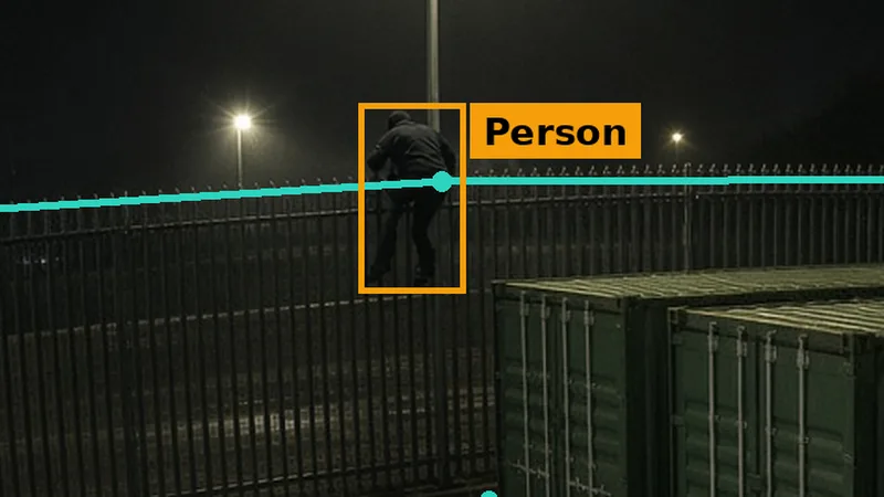 Phone alert crop: a figure on top of a storage yard palisade fence at night, crossing a teal fence line