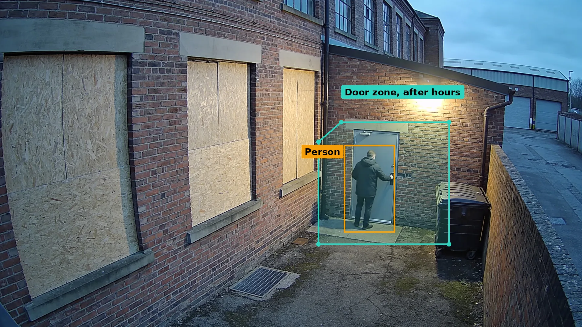 Dusk CCTV of a closed brick factory with boarded windows, a person at the rear door boxed in amber in a teal zone