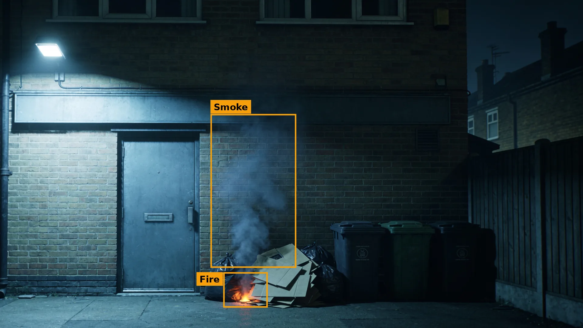 Night CCTV of the rear of an empty shop, bags and cardboard alight by the door, smoke and fire boxed in amber
