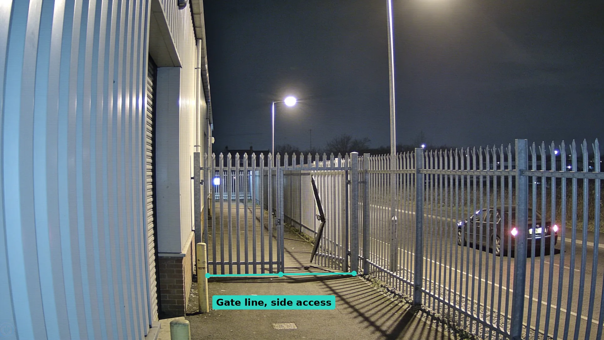 Night CCTV of a side gate beside an empty unit with one bent pale, a teal line drawn along the foot of the gate