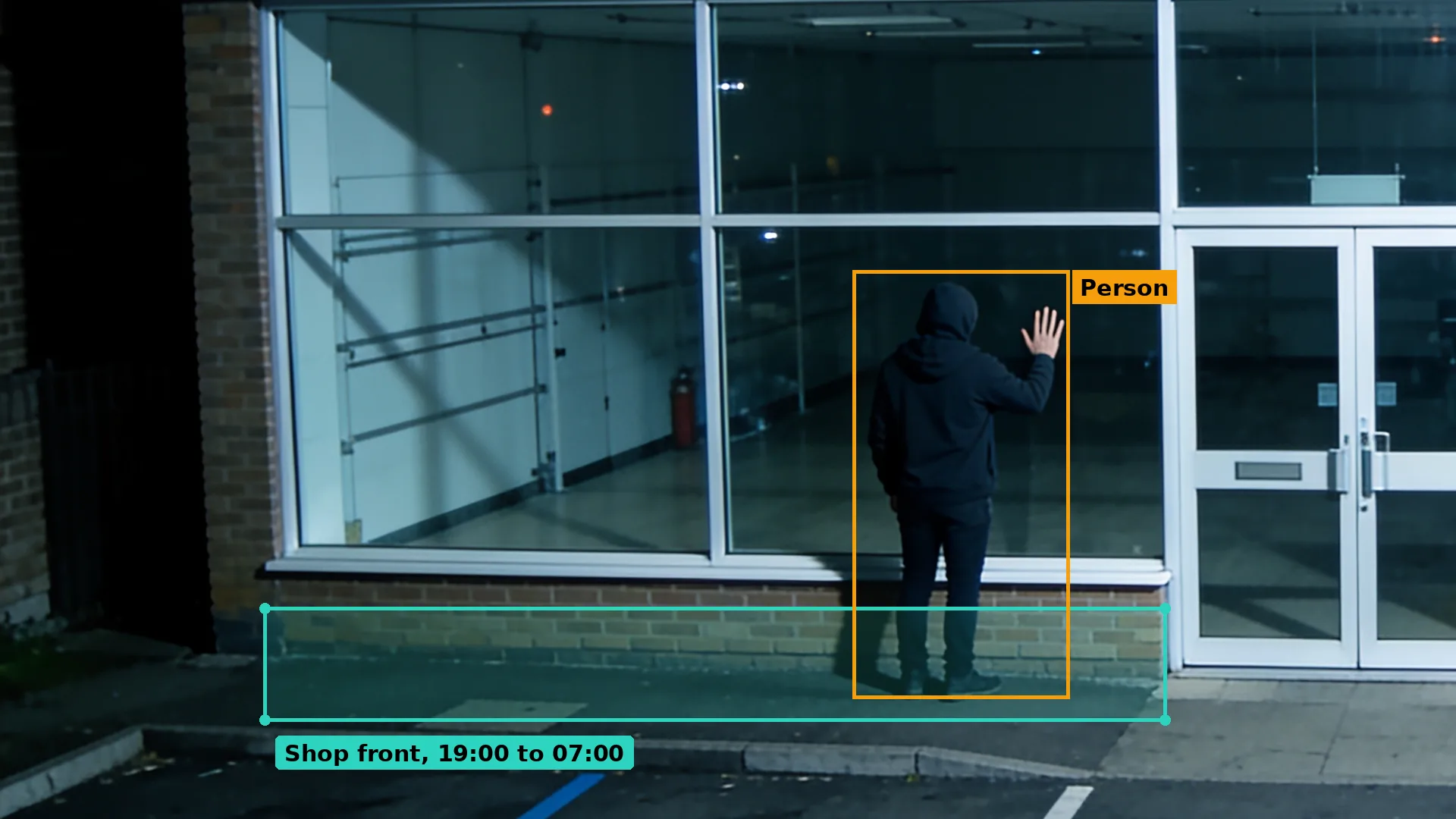 Night CCTV of a hooded person with a hand on the plate glass window of a closed trade counter, boxed in amber