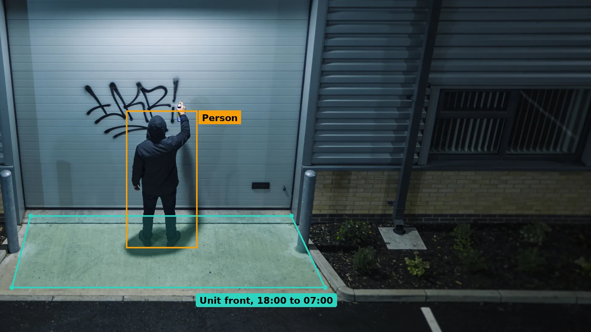 Night CCTV of a hooded person spraying a tag on the roller shutter of an industrial unit, boxed in amber