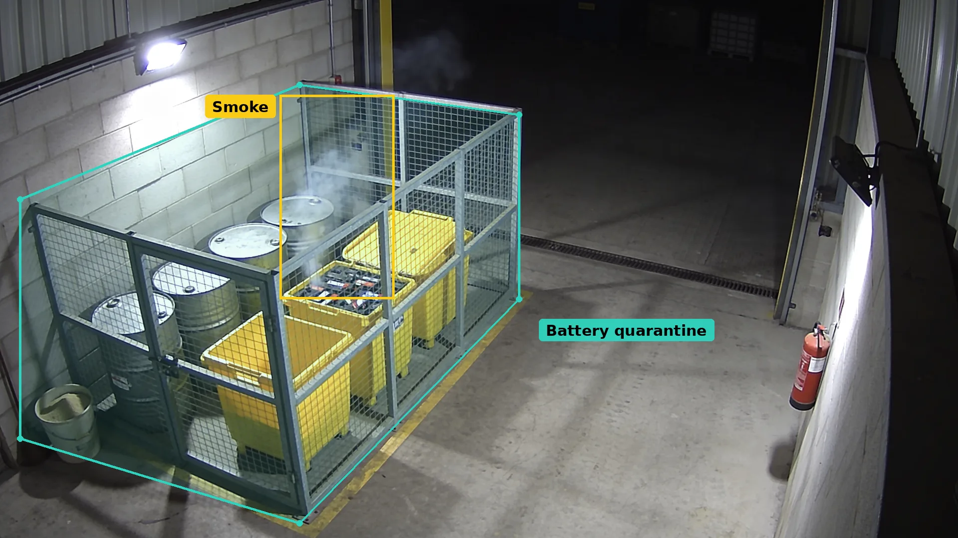 Night CCTV of a battery quarantine cage with yellow bins and drums in a teal zone, smoke boxed in amber