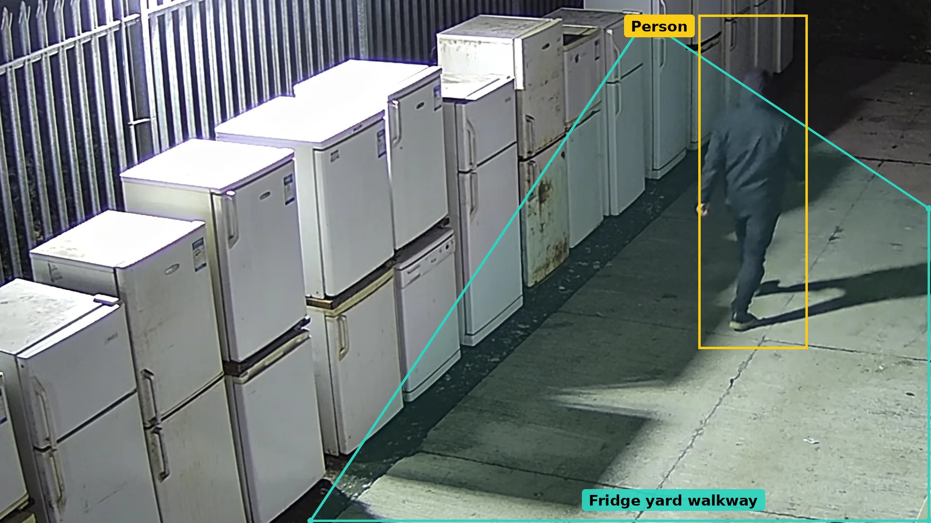 Night CCTV of a yard of stored fridges, the walkway in a teal zone and a person on foot boxed in amber