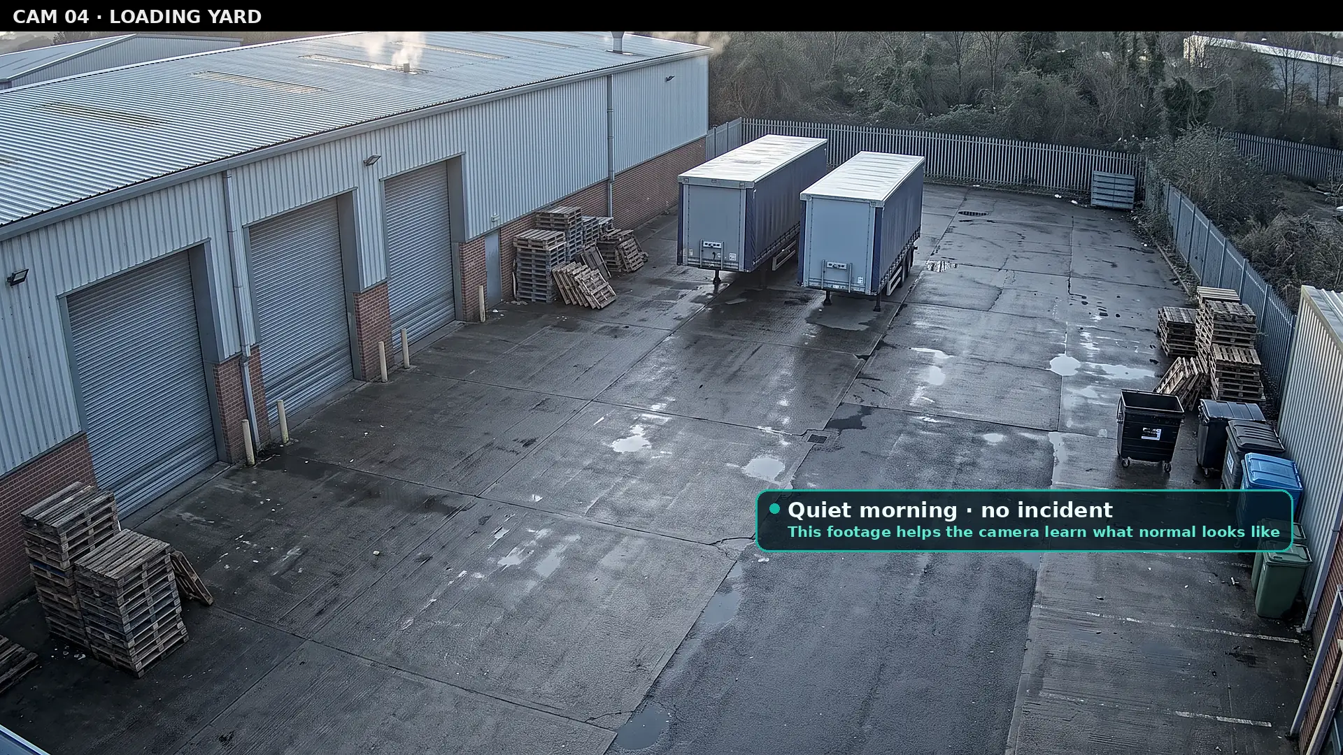 Elevated camera view of an empty loading yard in the early morning with closed roller shutters, two parked trailers, stacked pallets and bins, labelled as a quiet morning with no incident used to learn what normal looks like