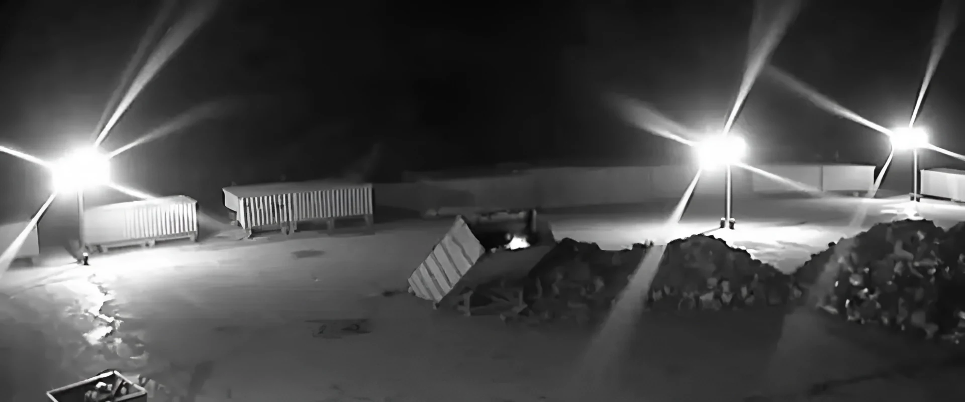 Real night-time infrared CCTV frame of a storage yard with floodlights, containers and stockpiles, with a small fire in a tipped skip