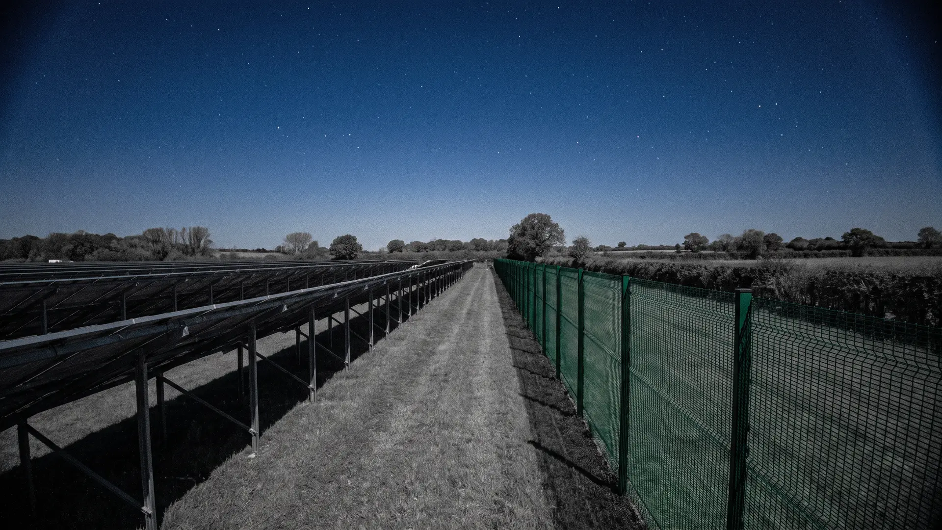 Night CCTV view: solar panel rows beside a mesh perimeter fence on a solar farm