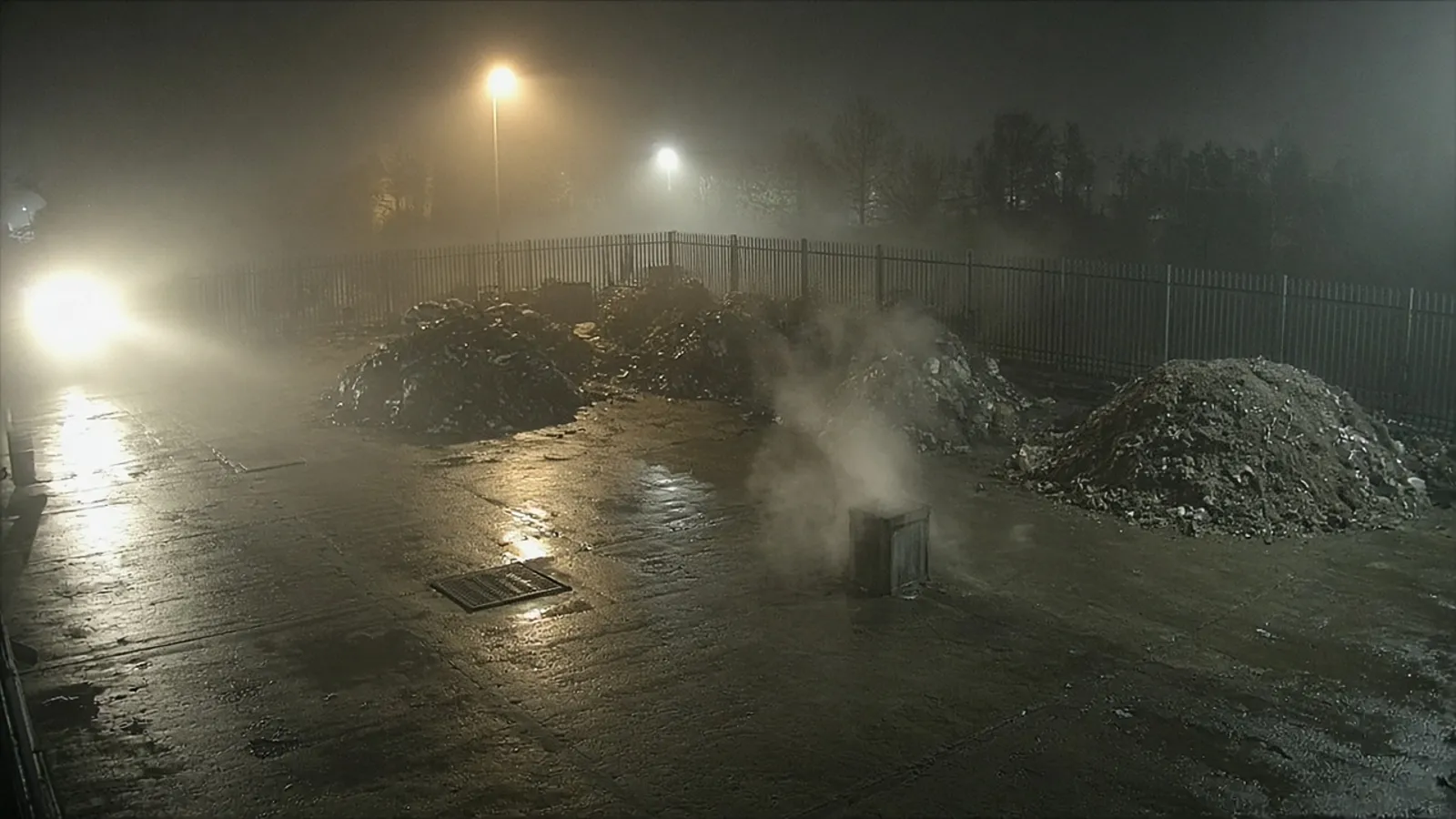 Illustrative image: night-time CCTV-style view of an empty yard with fog, steam rising from a vent and vehicle headlight glare across wet concrete