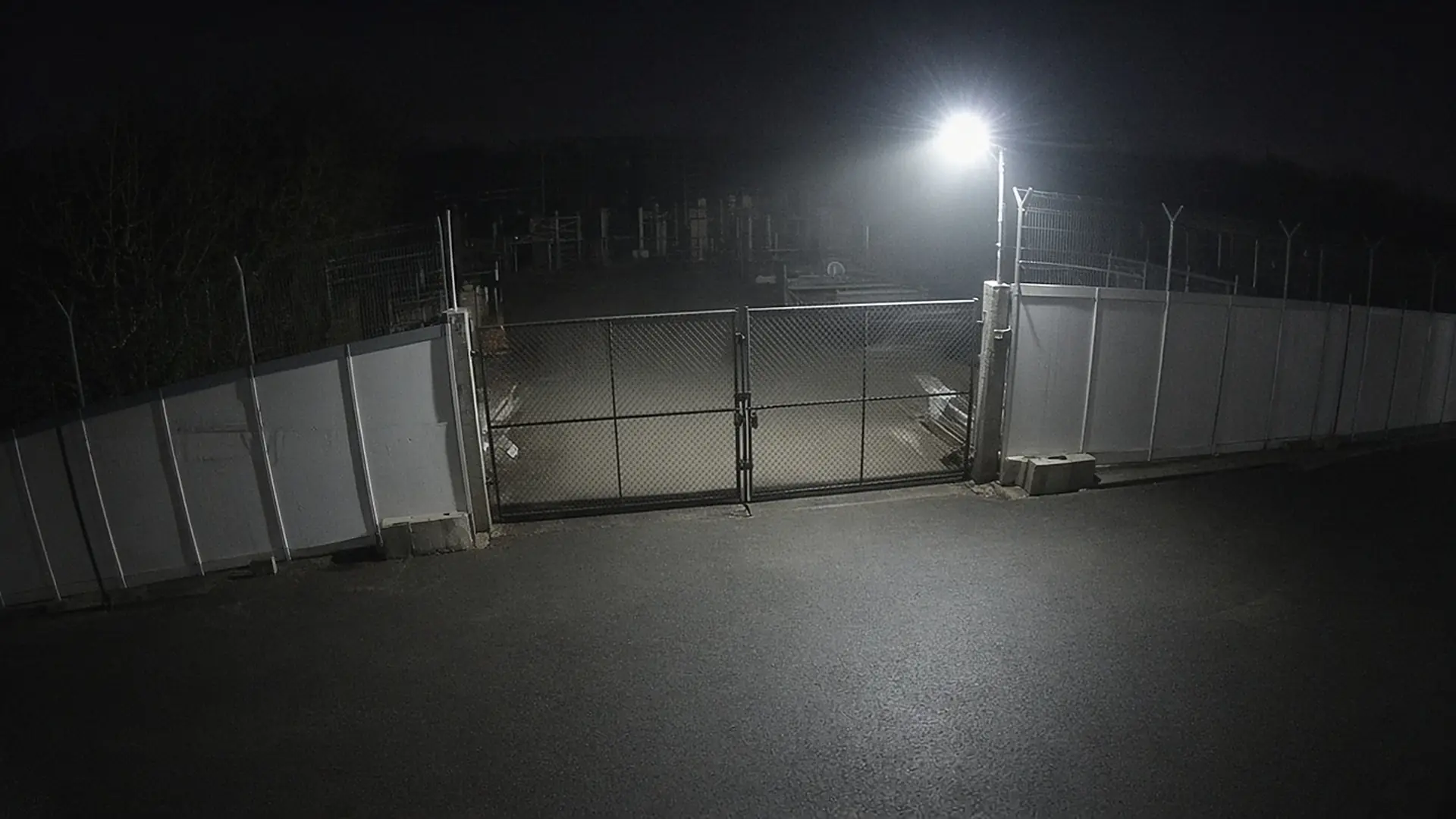 Night view of a closed mesh double gate between white hoarding panels under a floodlight, with nobody and no vehicles in view