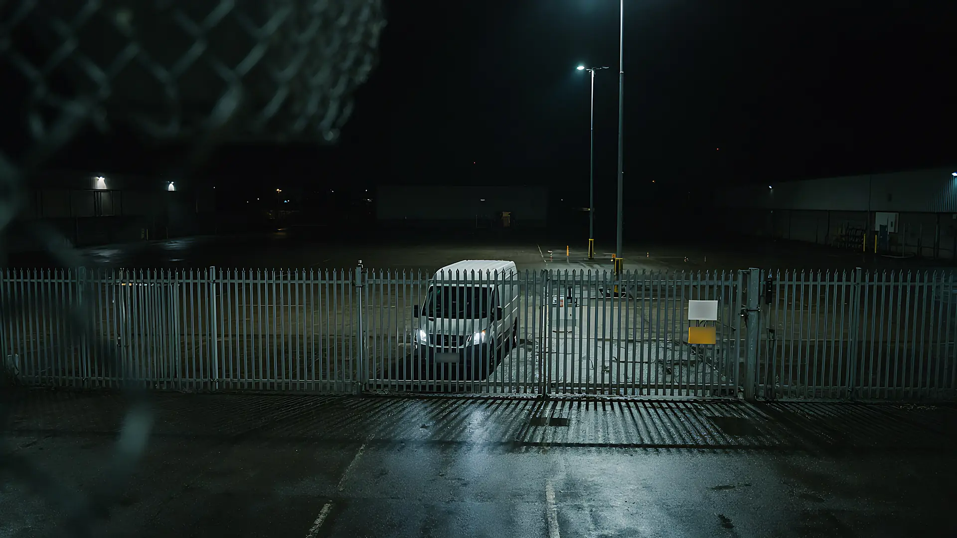 Night view through a palisade fence and closed gate into a yard, with a white van parked inside behind the fence