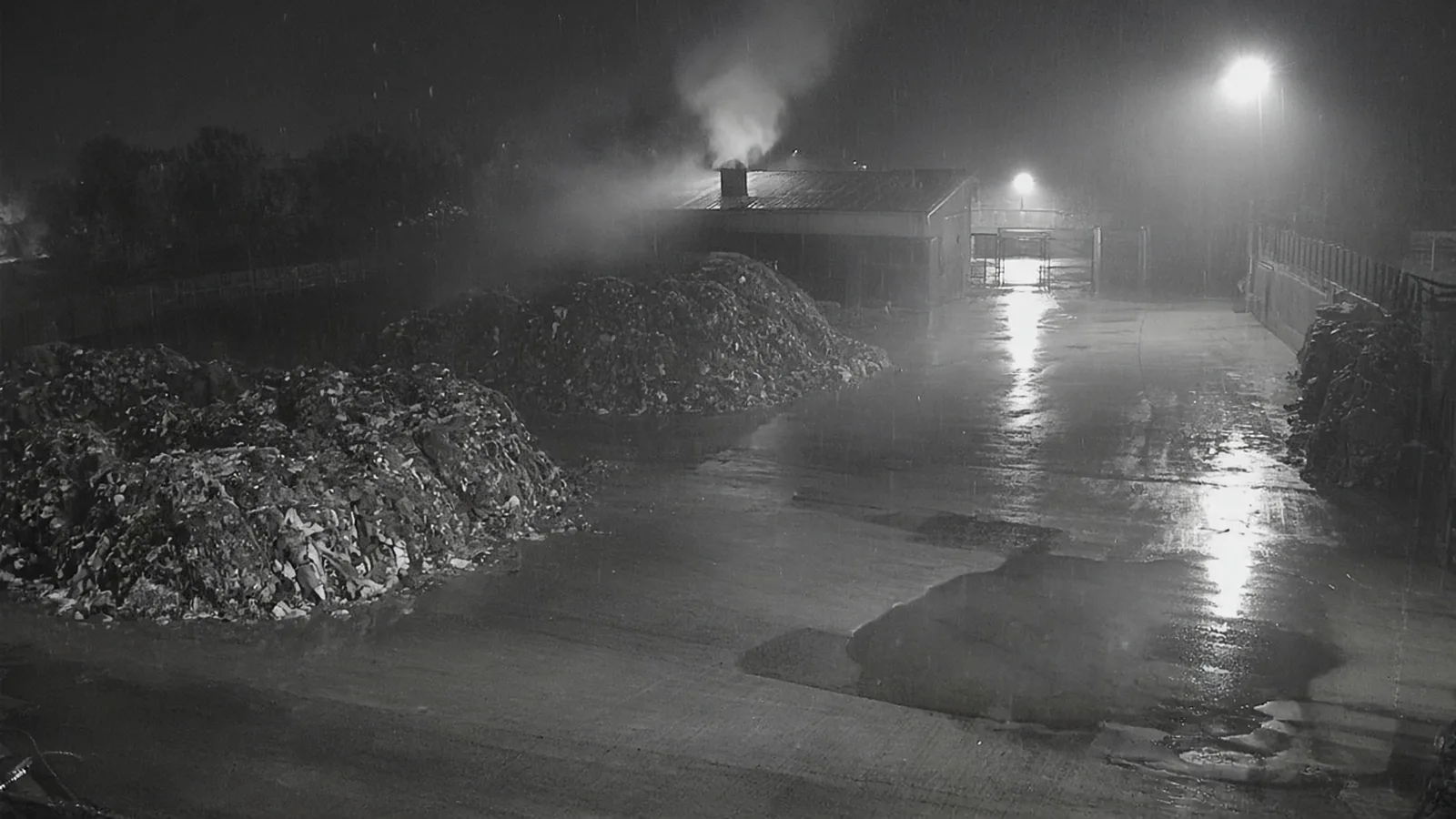 Illustrative night-time infrared CCTV view of an empty waste yard in rain and mist, with wet concrete reflecting floodlights, two stockpiles and steam rising from a vent on a shed roof