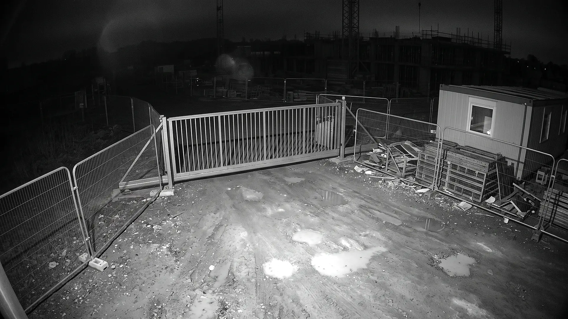 Night-time infrared camera view of an empty site gate in black and white, with the turnstile and fencing lit by infrared